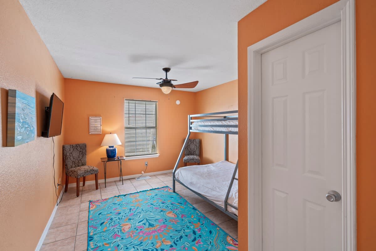 Vibrantly decorated bedroom with bright orange walls, a metal bunk bed, and a colorful blue and pink floral rug. A wall-mounted TV and a window with blinds are also visible.