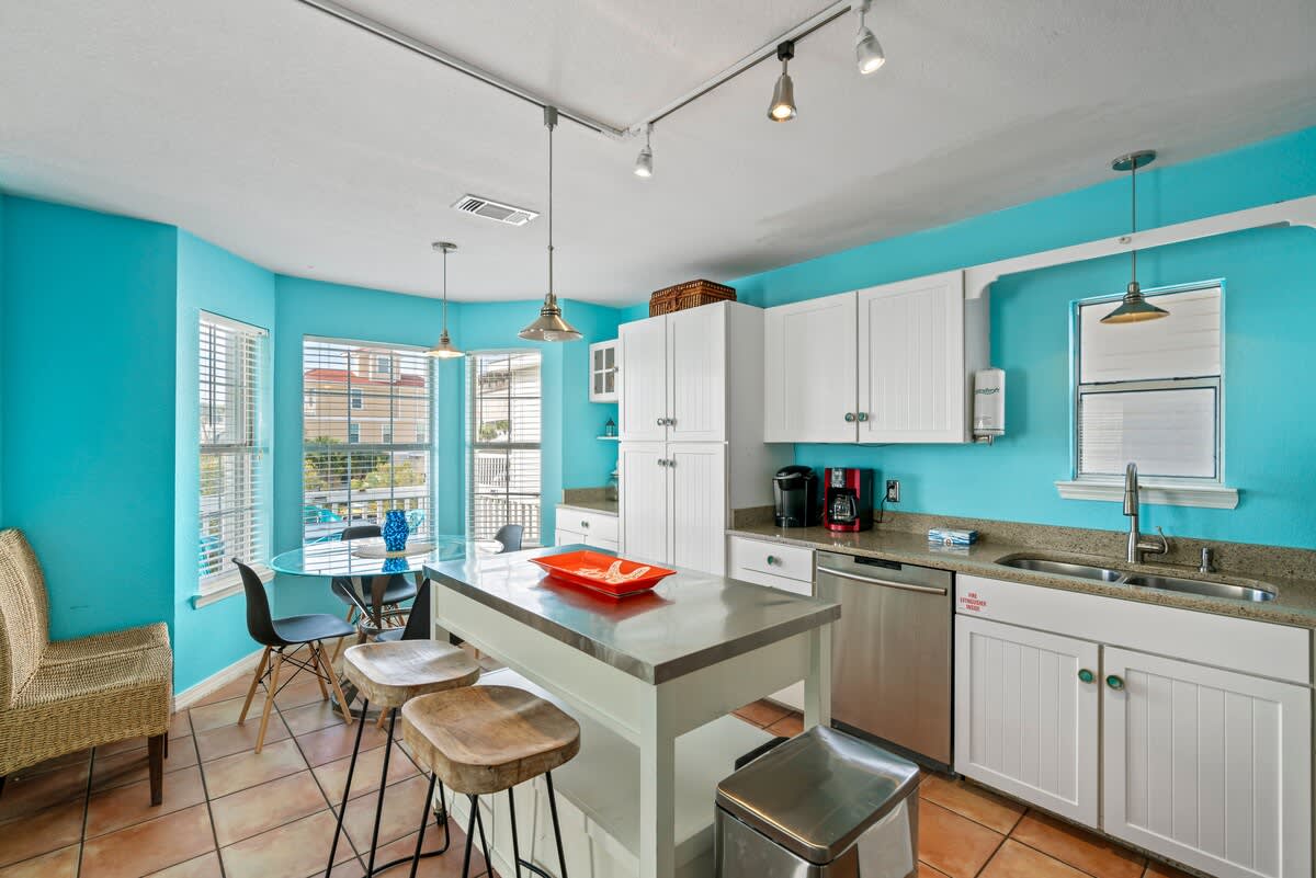 Vibrant kitchen and dining area with bright blue walls, white shaker cabinets, stainless steel appliances, and a central island with bar stools, adjacent to a dining table by large windows.