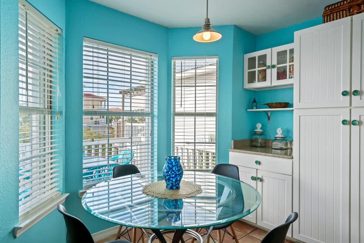 Bright dining nook with a round glass table and black chairs, surrounded by multiple windows with blinds, built-in white cabinets, and vibrant turquoise walls, offering a view of the outdoors.