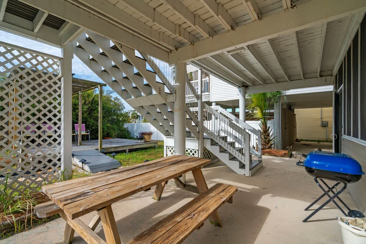 A concrete patio area beneath the house features a rustic wooden picnic table and a blue BBQ grill, with white wooden stairs leading up to the main level and a lattice fence enclosing part of the space.