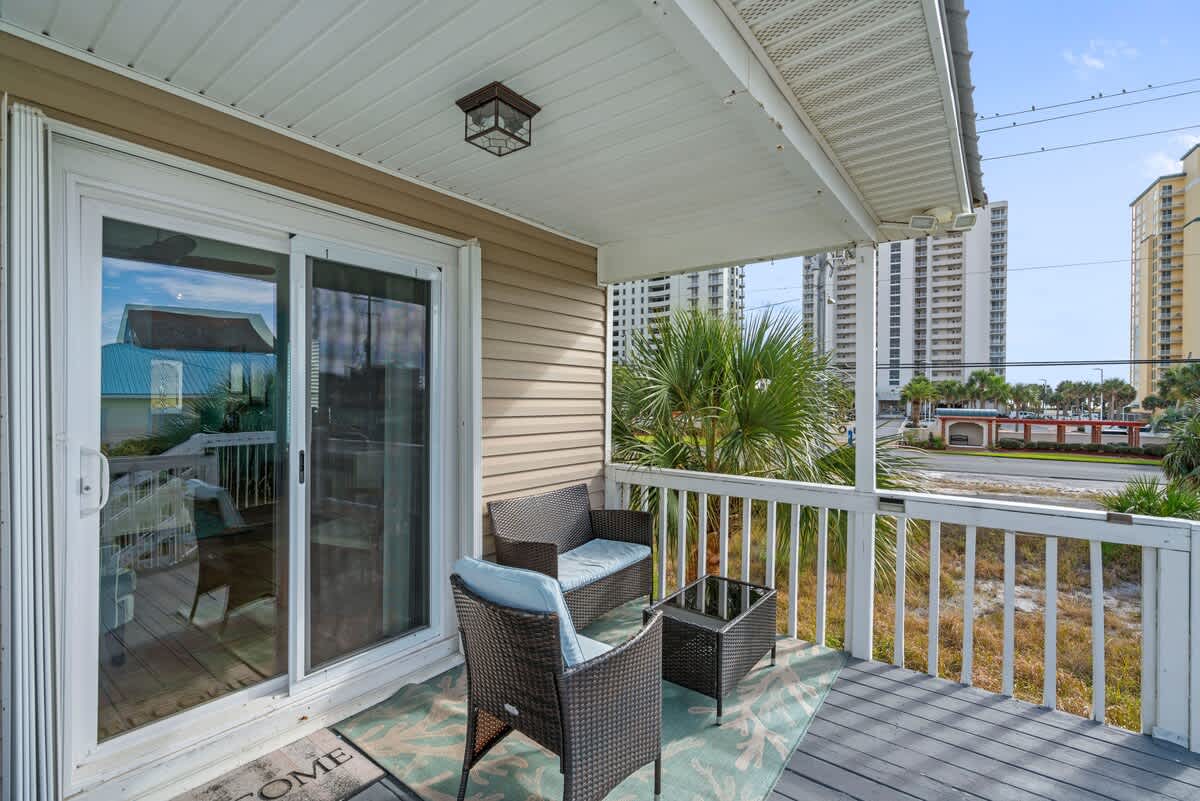 An elevated balcony features a small outdoor seating arrangement with two wicker chairs and a matching side table, set on a blue and white patterned rug, overlooking a street and distant high-rise buildings.