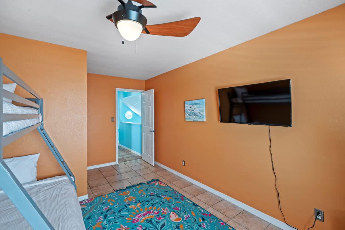 Another angle of the vibrant bedroom with orange walls, showcasing a metal bunk bed, a wall-mounted TV, and a colorful floral rug. A doorway leads to another room with blue walls.