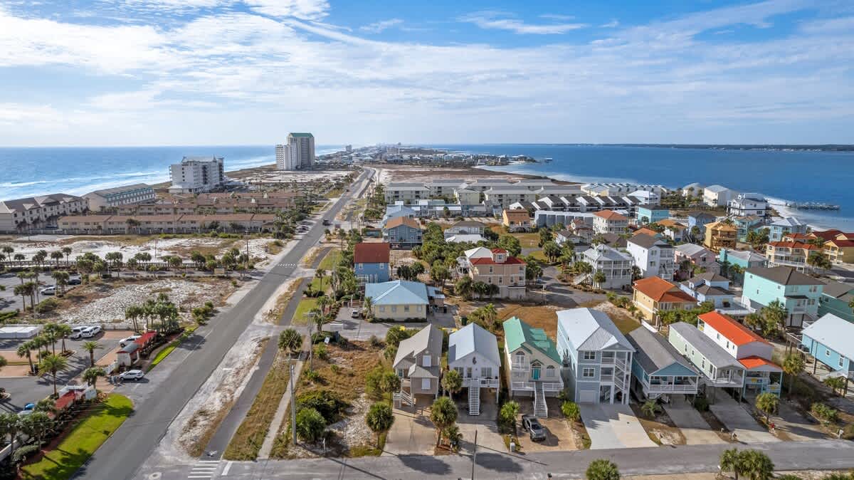 Panoramic aerial view of a coastal community with numerous colorful houses, a main road, and both the ocean and a bay visible in the distance under a bright blue sky.