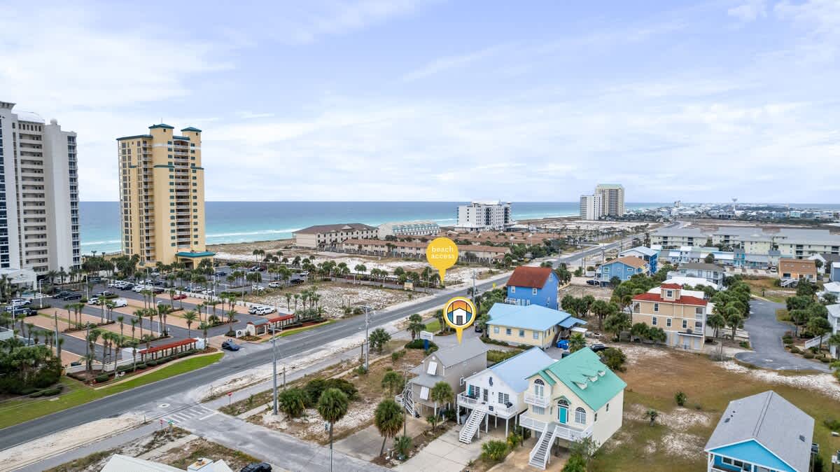 A wide aerial shot captures the property, marked by a house icon, nestled among other homes on a street, with the expansive ocean and distant high-rise buildings forming the backdrop, and a 'beach walkway' pin indicating nearby access.
