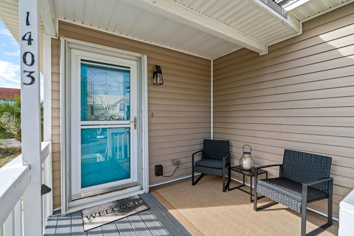 Inviting front porch entrance with light brown siding, a white screen door, and a 'WELCOME' mat. Two dark wicker chairs and a small table create a cozy seating area.
