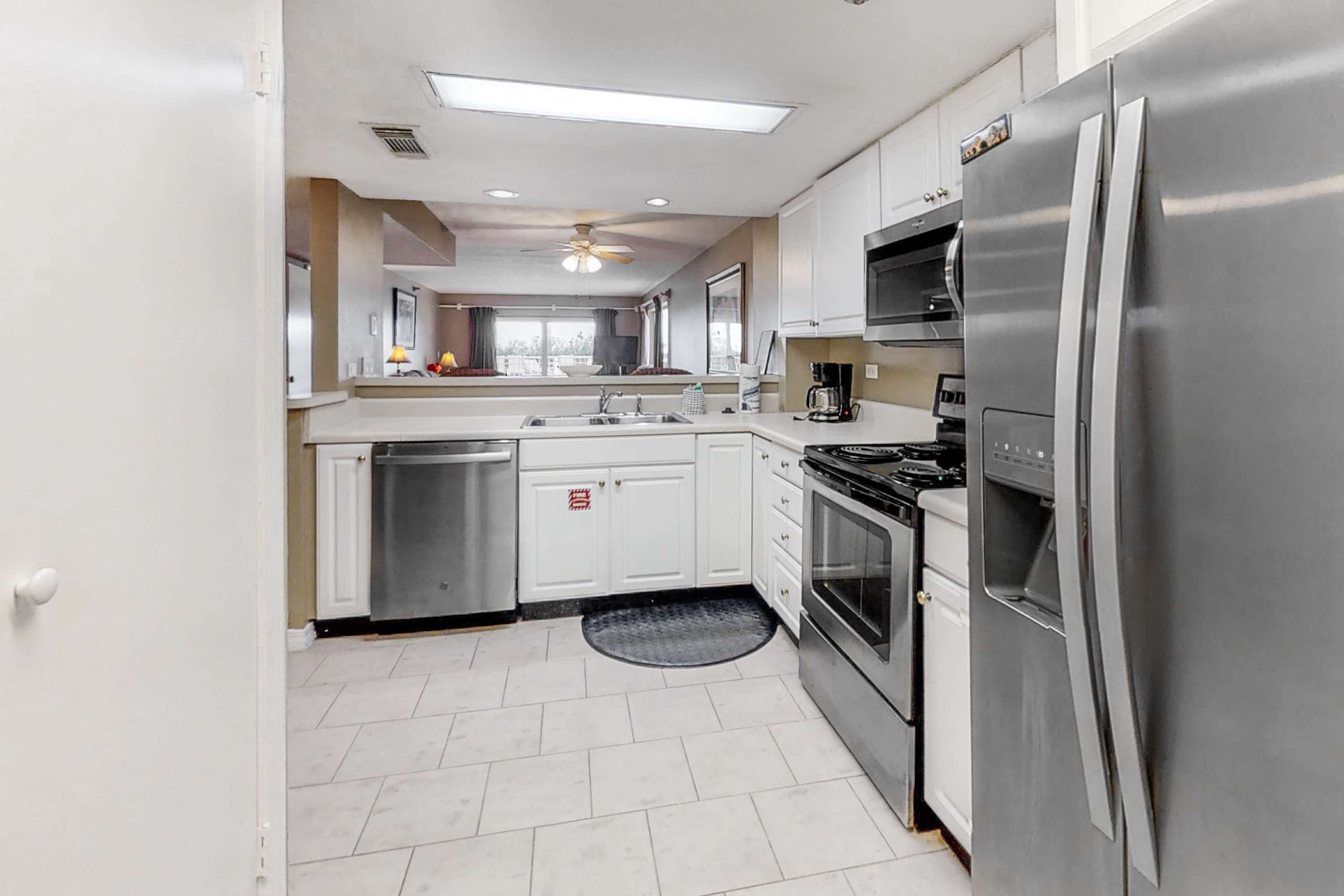 Wider view of the kitchen, showing white cabinets, stainless steel appliances, a double sink, and a pass-through window looking into an adjacent living area.