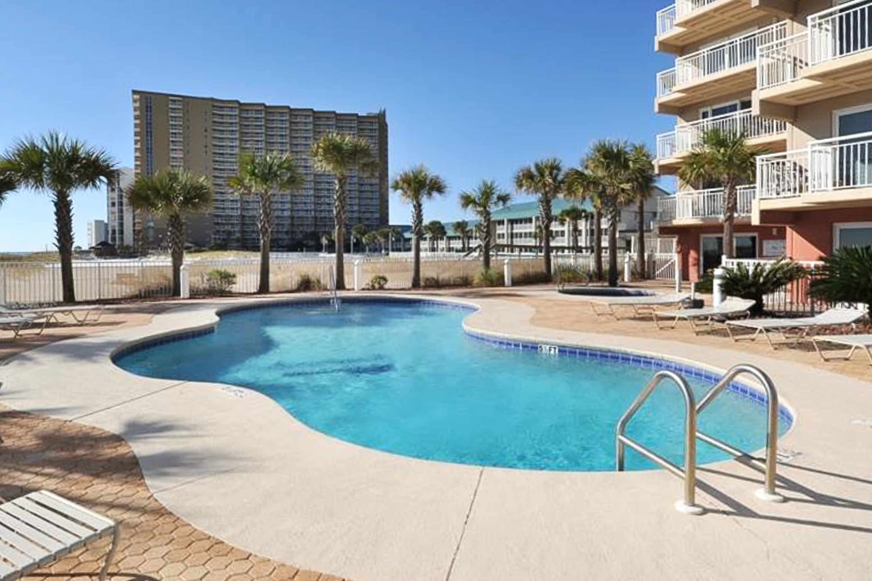 An outdoor communal pool with a freeform shape is surrounded by a light-colored deck and several lounge chairs, with palm trees and a tall building in the background under a clear blue sky.