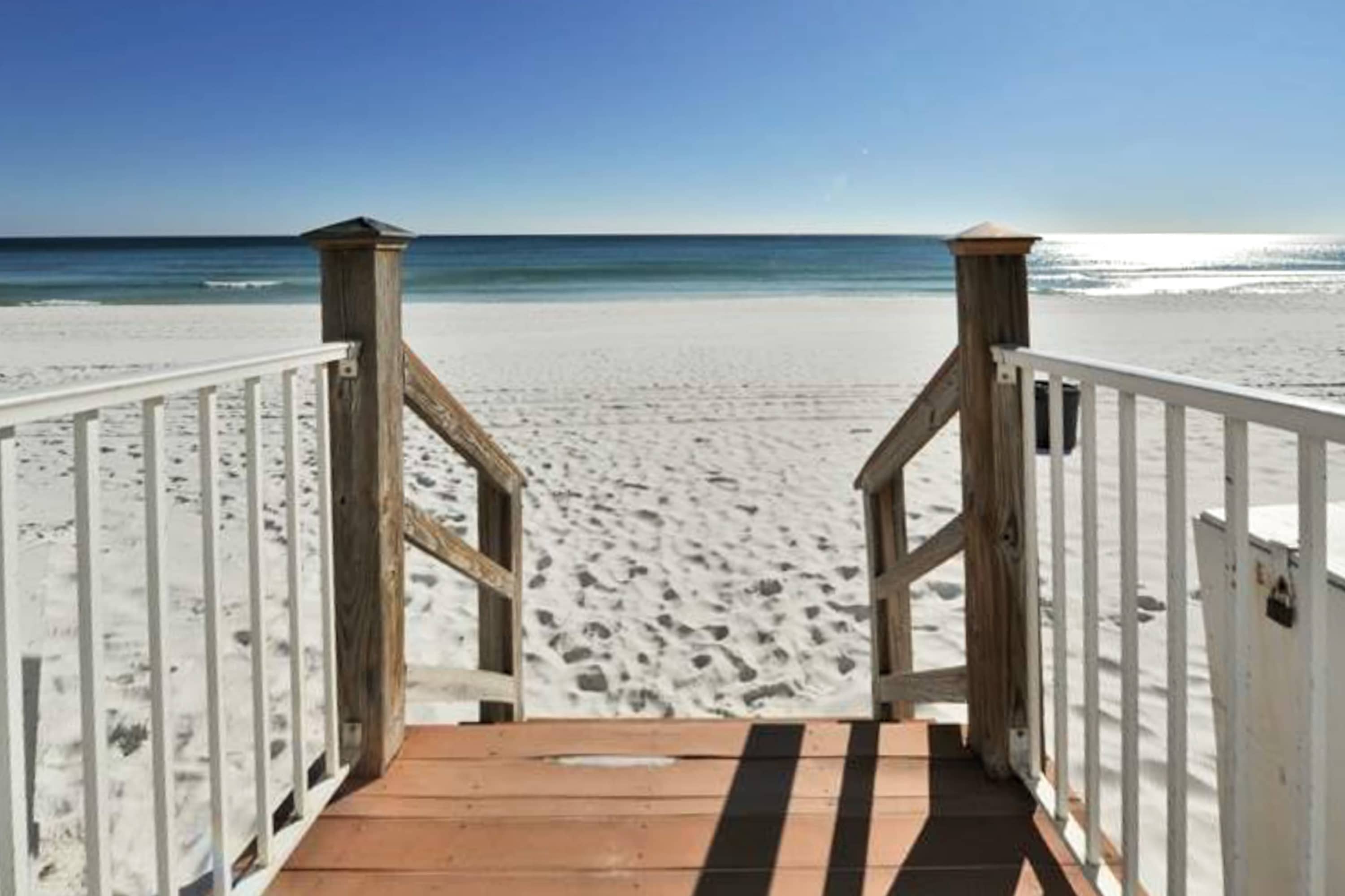 A wooden walkway with white railings leads down to a wide, pristine white sand beach and the sparkling blue ocean under a bright sunny sky.