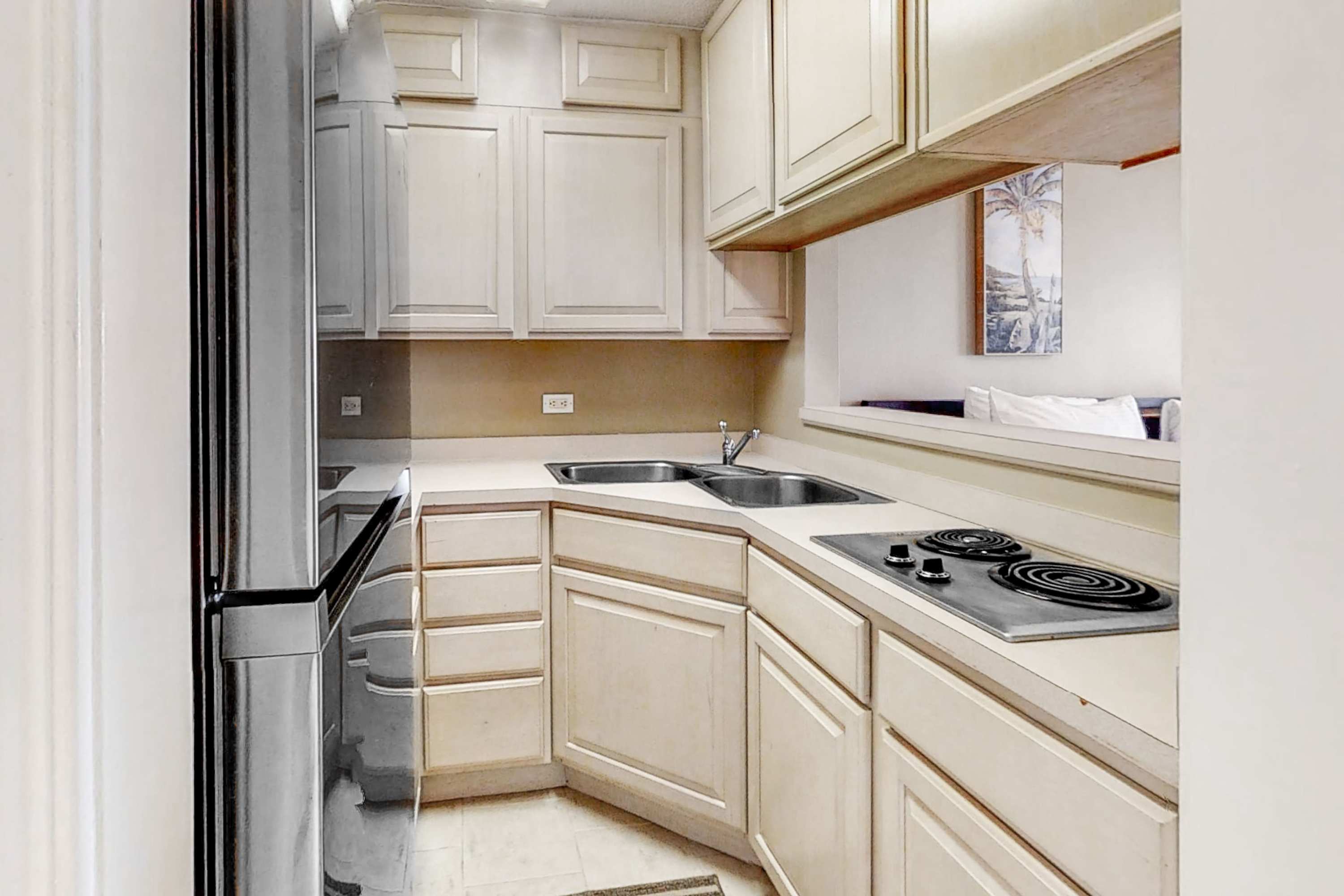 A compact kitchen features light cream cabinetry, a stainless steel refrigerator, a double sink, and a black electric stovetop set into a light-colored countertop. A small pass-through window looks into an adjacent living area.