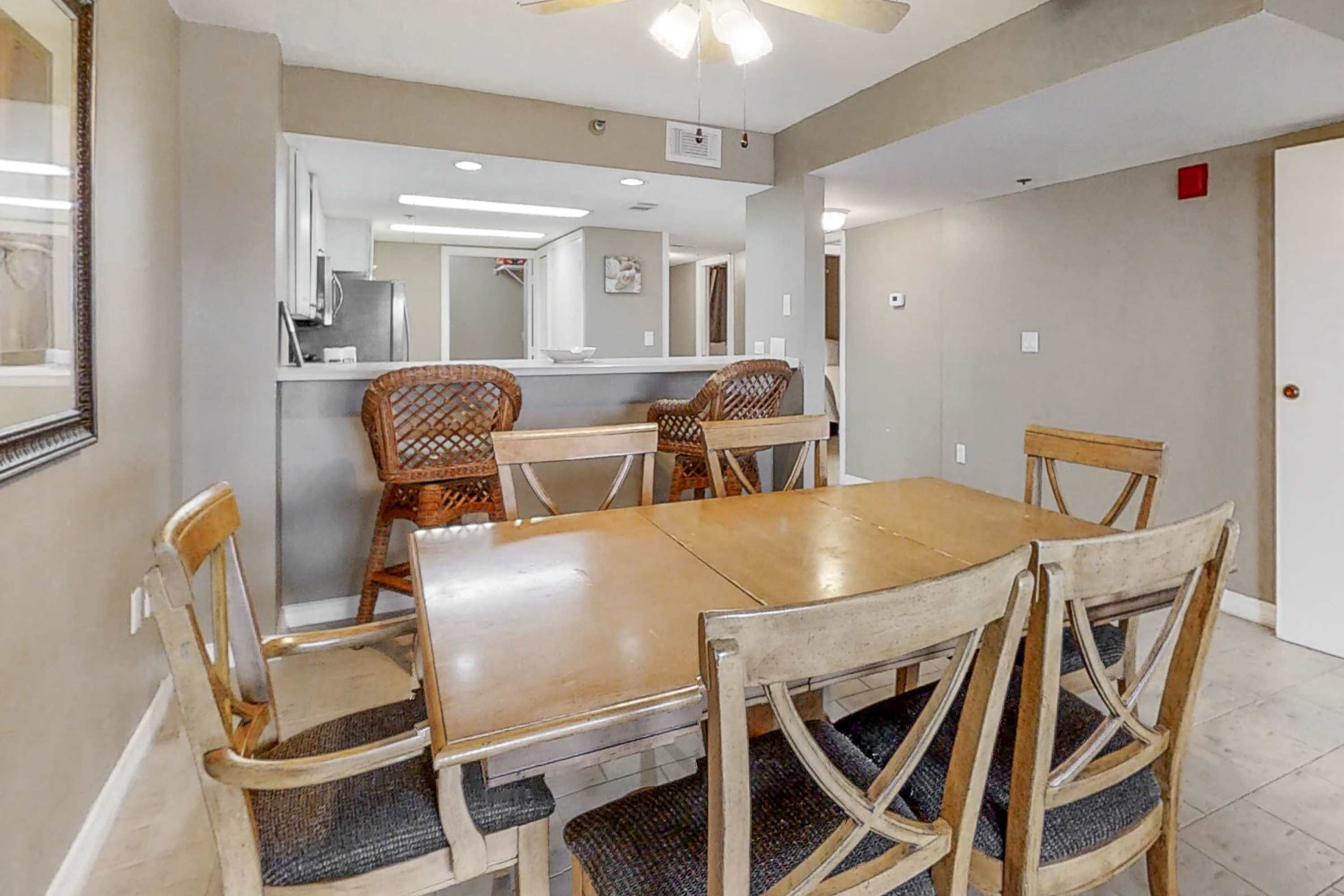 A closer view of the dining area, showcasing the wooden dining table and chairs with patterned cushions. The breakfast bar with two wicker stools provides a clear view into the white-cabinet kitchen with stainless steel appliances.