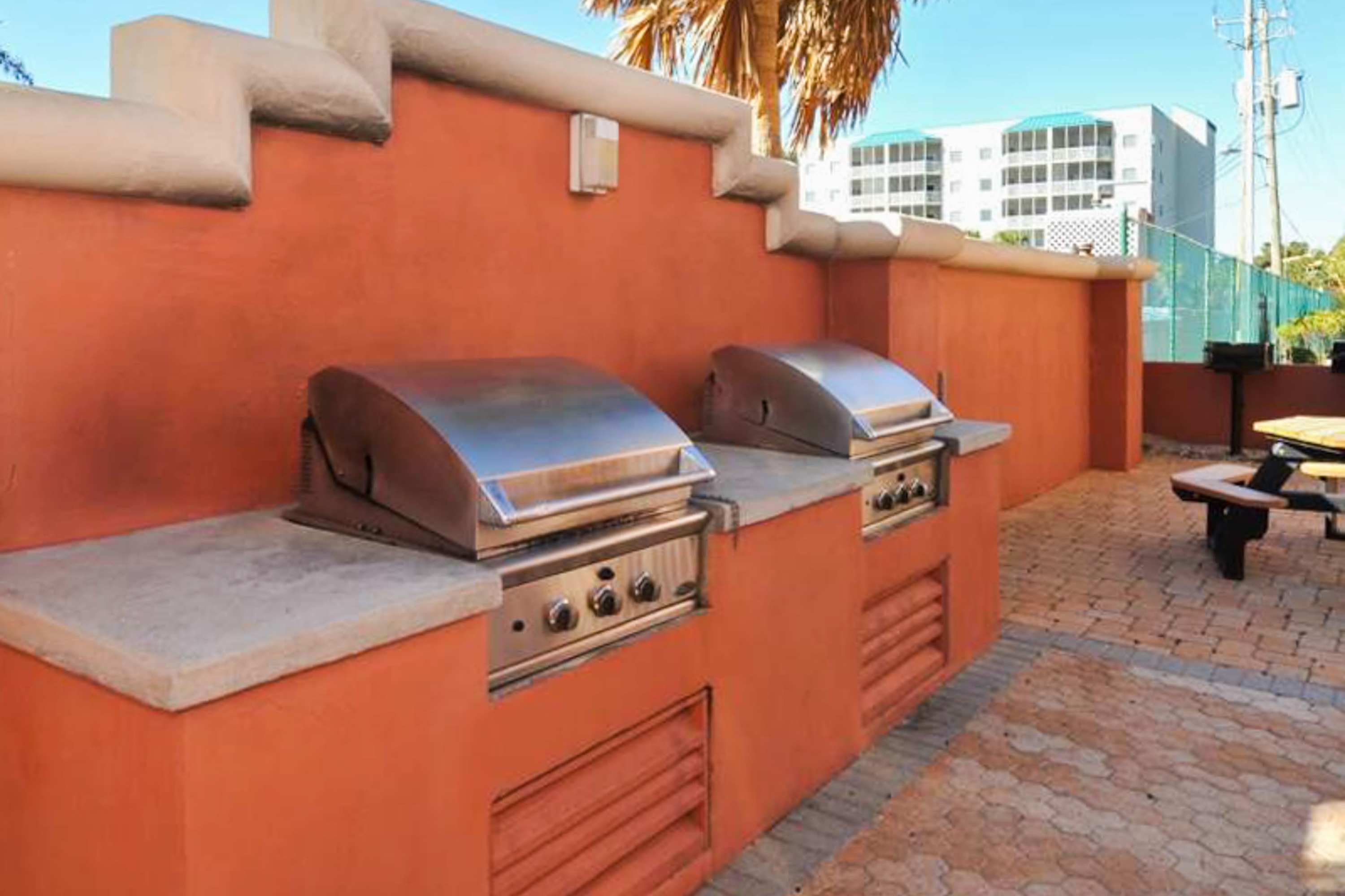 Two built-in stainless steel BBQ grills are set into an orange stucco structure on a paved patio, with a picnic table visible in the background.