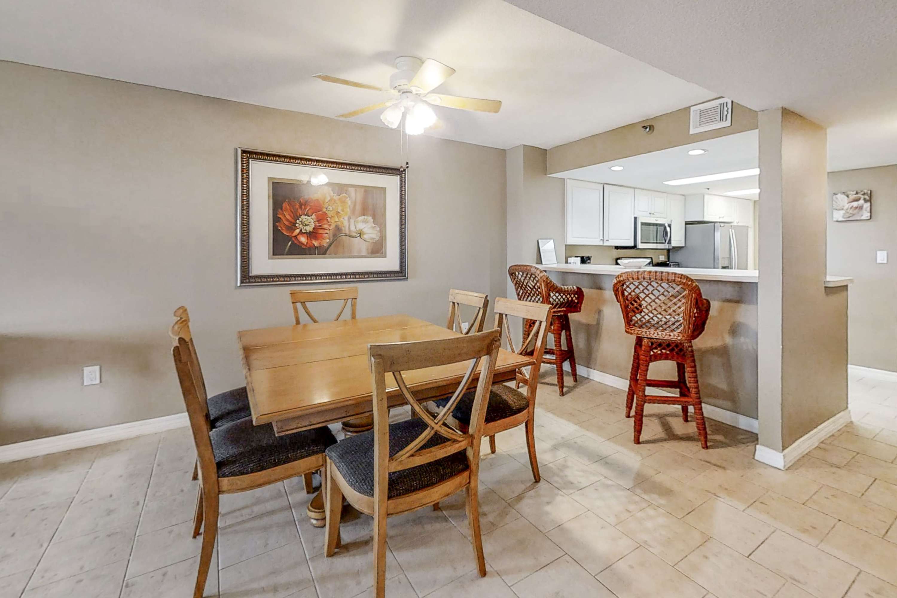 A warm dining area featuring a wooden rectangular dining table with six chairs, set on light tile flooring. A large framed floral artwork hangs on the wall, and a breakfast bar with two wicker stools connects to the kitchen in the background.