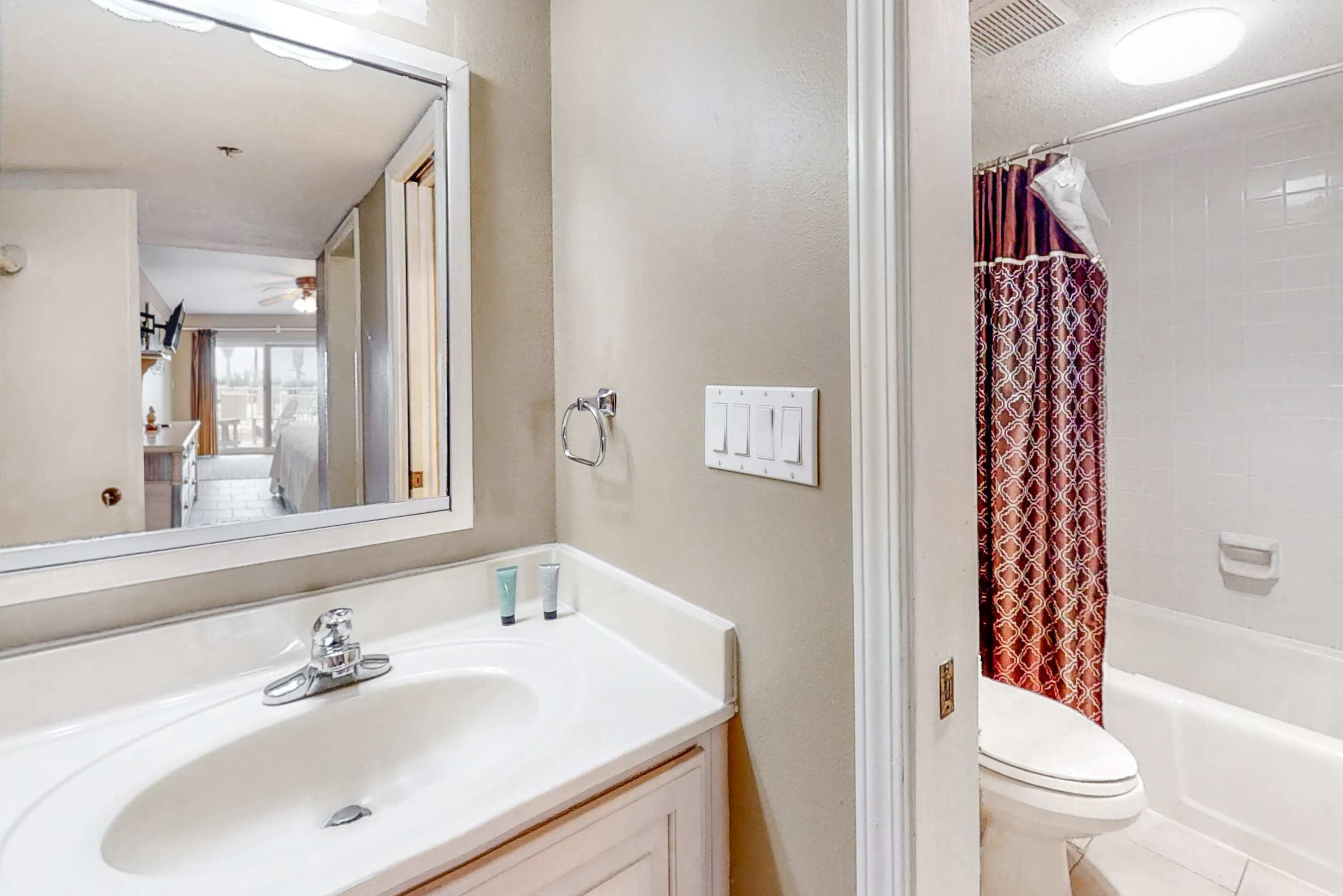A bright bathroom features a white vanity with a single sink, a large mirror, and a toilet next to a shower/tub combo with a patterned red and brown shower curtain. The walls are painted a light beige.