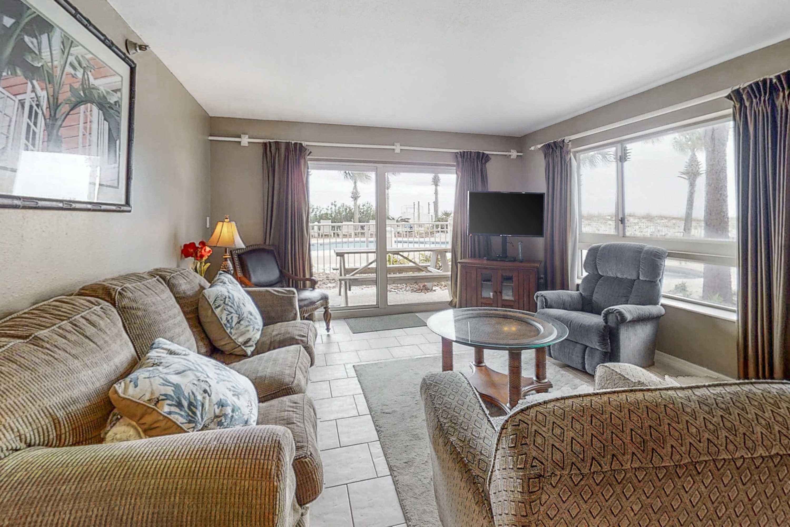 A cozy living room features a large brown sofa, a patterned armchair, and a glass-top coffee table on a rug, with a flat-screen TV and large windows offering views of the outdoor pool area and palm trees.