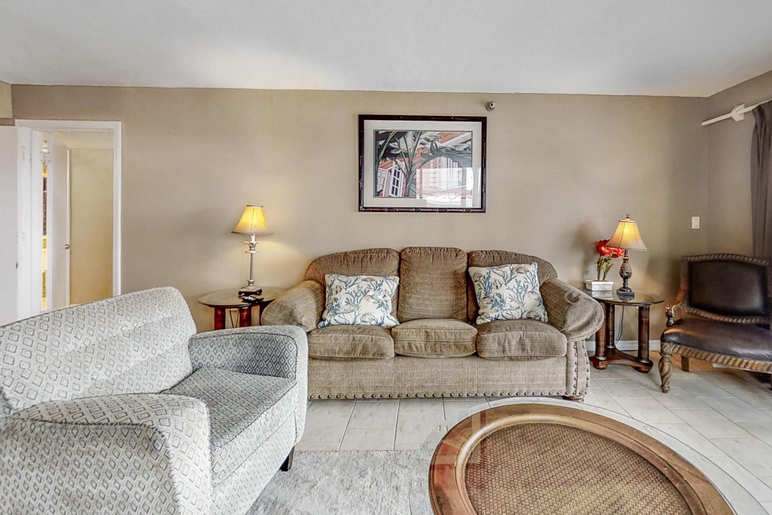 The living room is furnished with a long brown sofa adorned with decorative pillows, a light-patterned armchair, and two end tables with lamps, all set against a neutral wall with a framed artwork.