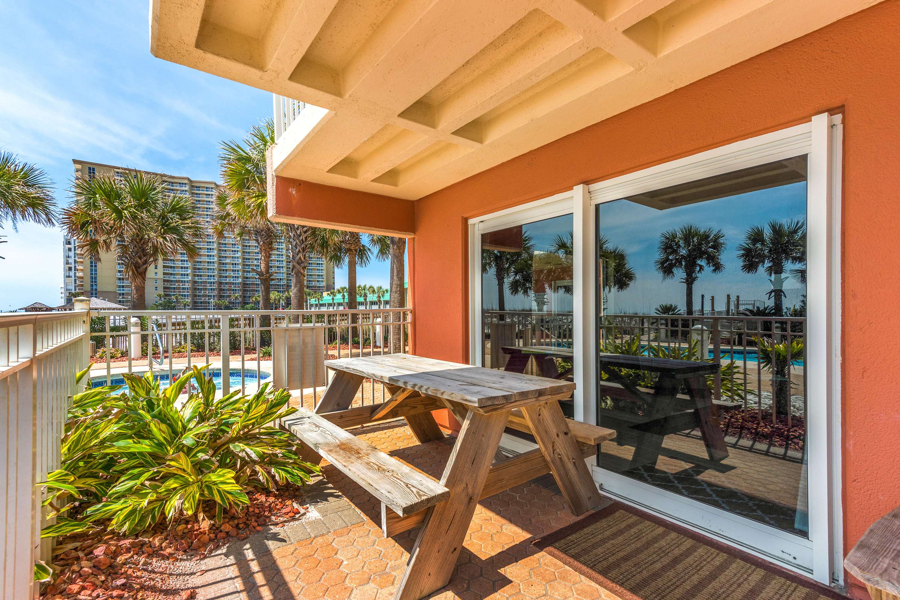 Spacious outdoor patio with a wooden picnic table, an orange exterior wall, and large sliding glass doors, offering views of the pool area and palm trees.