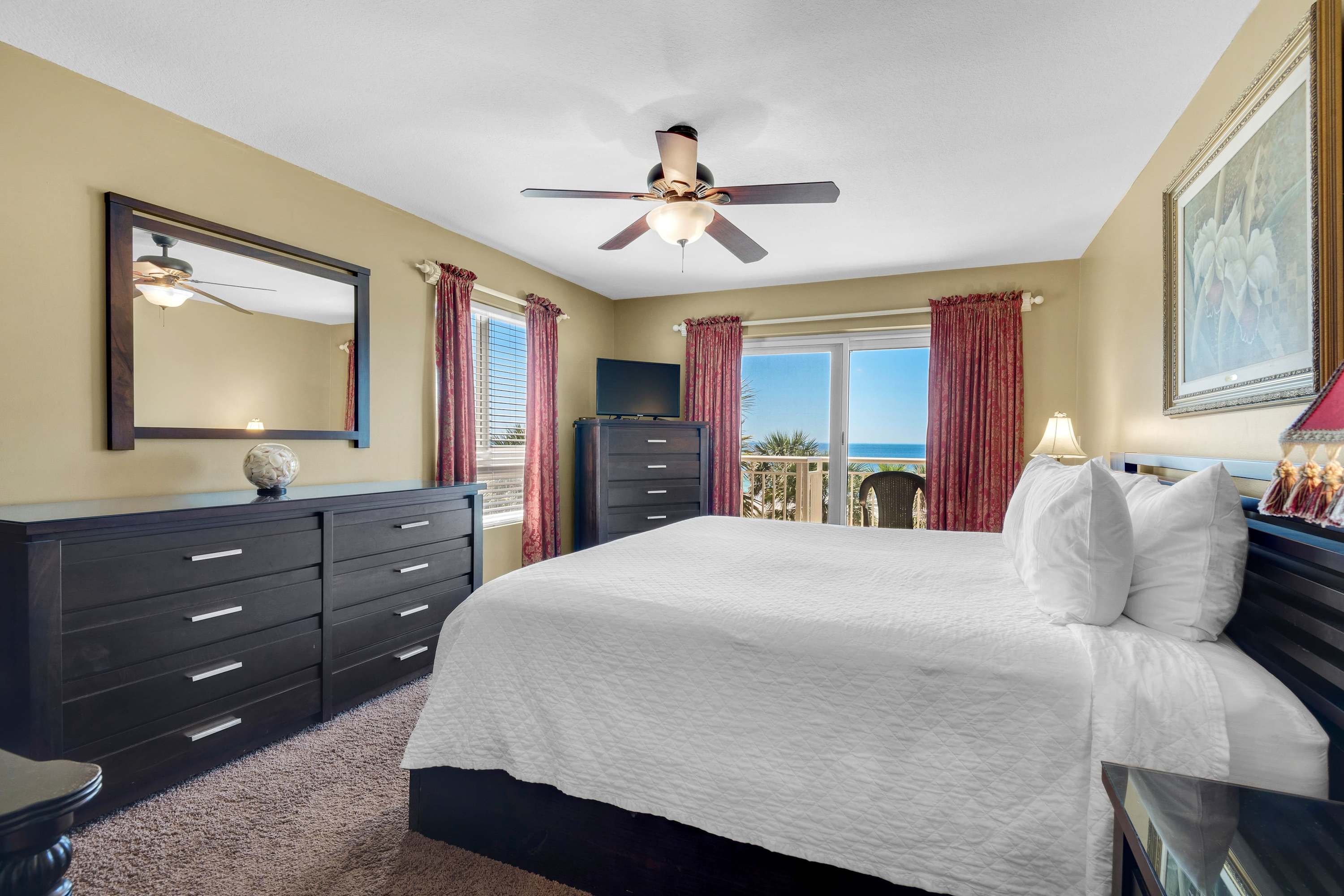 Master bedroom with a king bed, dark wood dresser, and a flat-screen TV, offering a bright view of the ocean and balcony through large windows with red curtains.