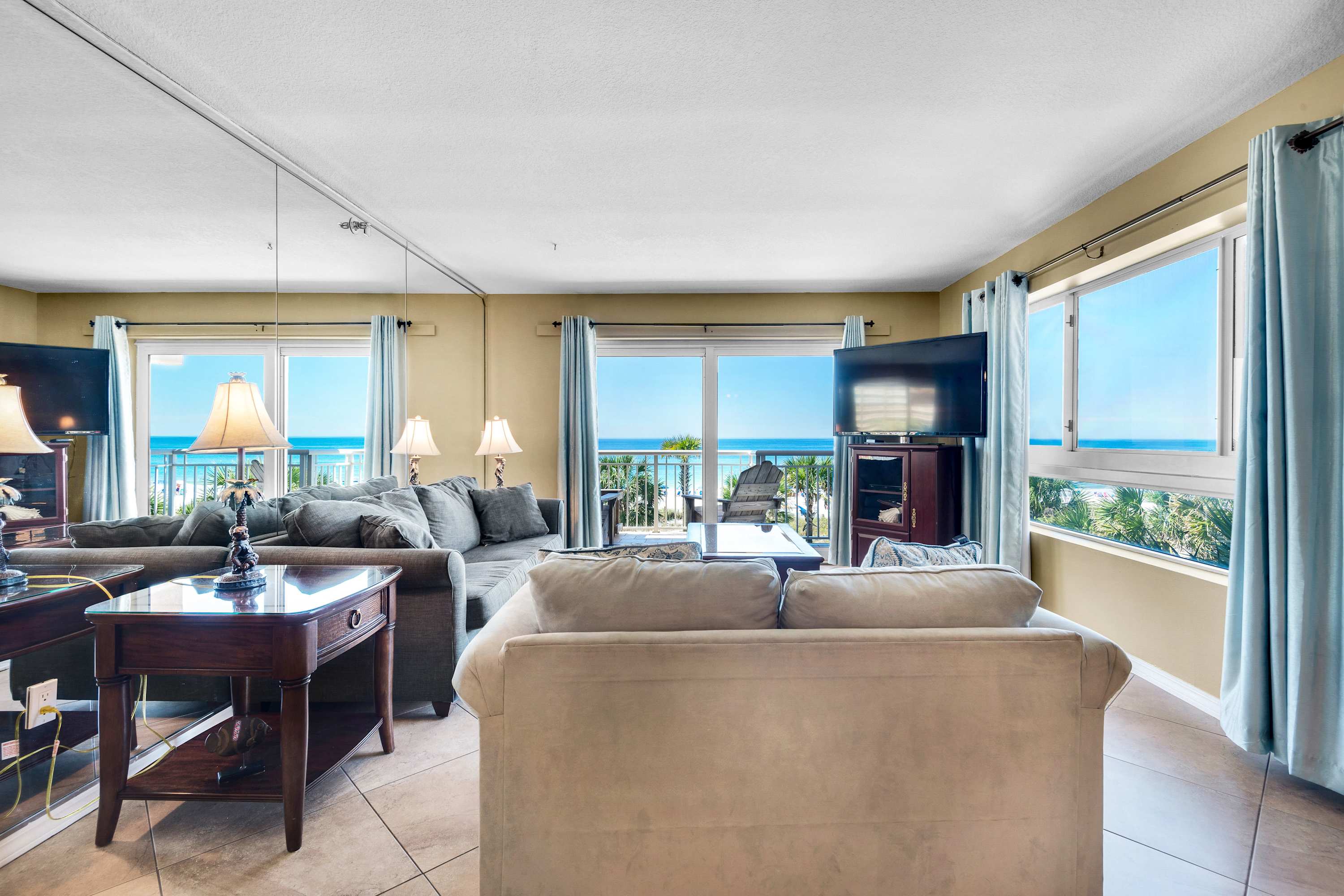 A bright living room with light-colored walls and tiled flooring, featuring a large gray sofa and a lighter-colored sofa facing a wall-mounted TV. Floor-to-ceiling windows and a sliding glass door offer panoramic ocean views and access to a balcony.