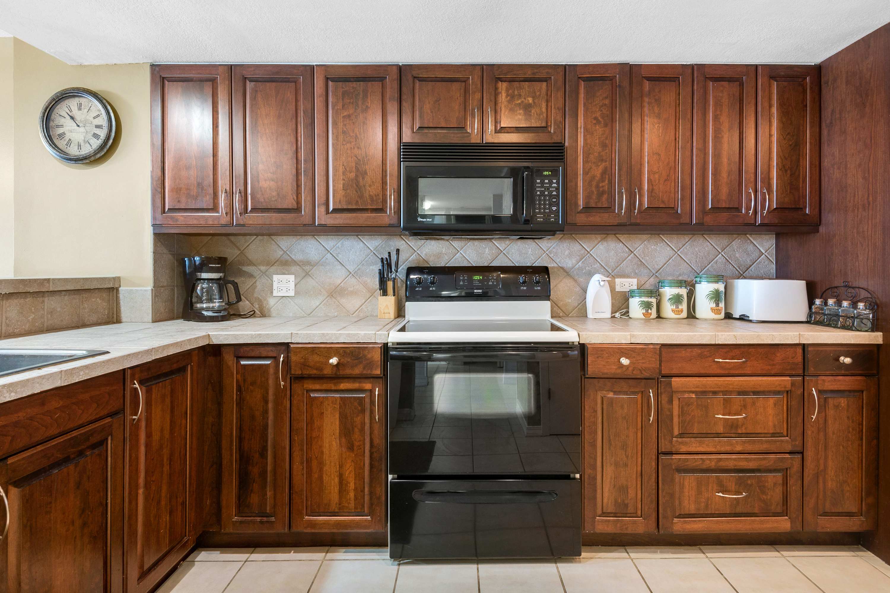 A close-up view of the kitchen counter shows dark wood cabinetry, light tiled countertops, and a black electric range with an overhead microwave. Various small appliances like a coffee maker, toaster, and decorative canisters are arranged on the counter.
