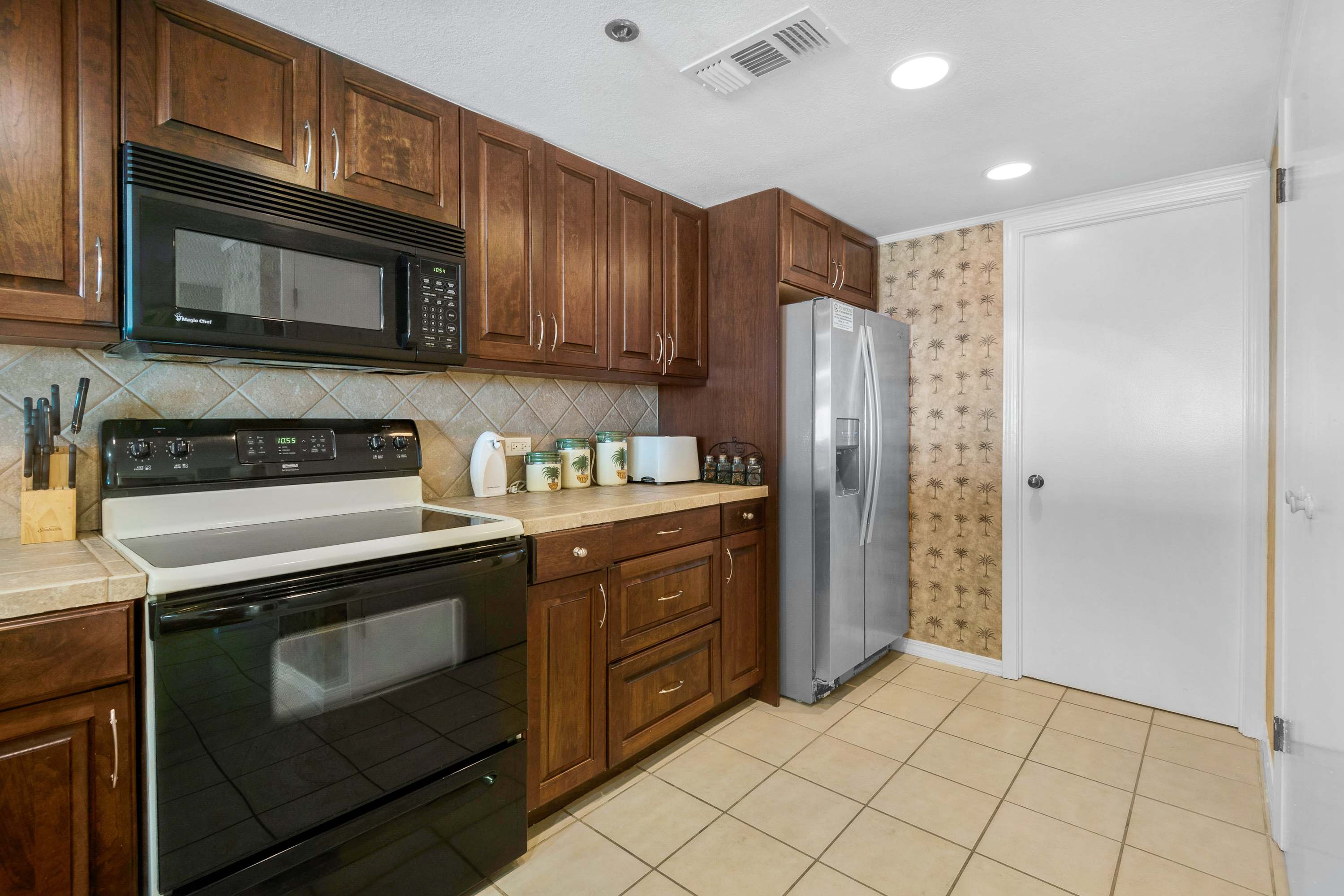 A well-lit kitchen features dark wood cabinetry, light-colored tiled countertops, and a black electric range with an overhead microwave. The walls are partially covered with a tropical-patterned wallpaper, and a stainless steel refrigerator stands at the end of the counter.