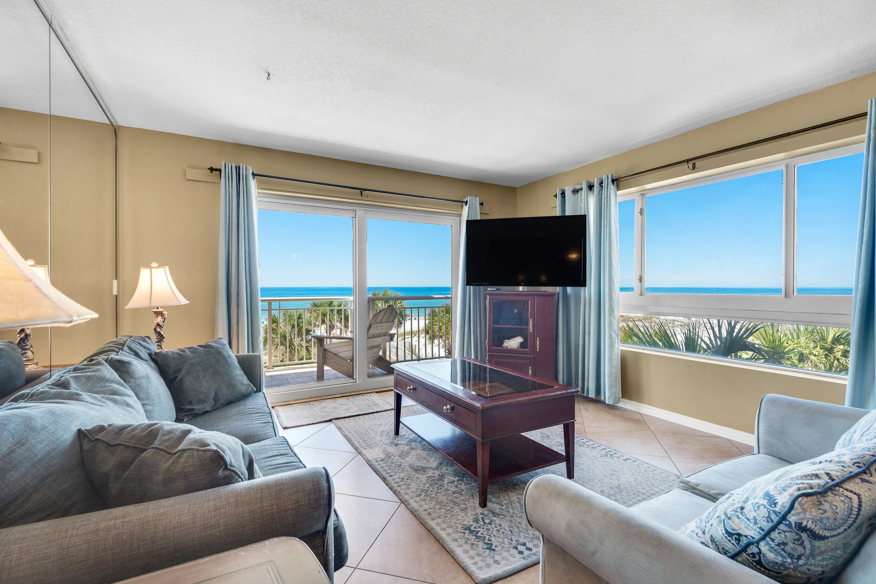 A comfortable living room with light-colored walls and tiled flooring, furnished with a gray sofa and a lighter sofa, centered around a dark wood coffee table. Large windows and a sliding glass door provide abundant natural light and stunning ocean views.