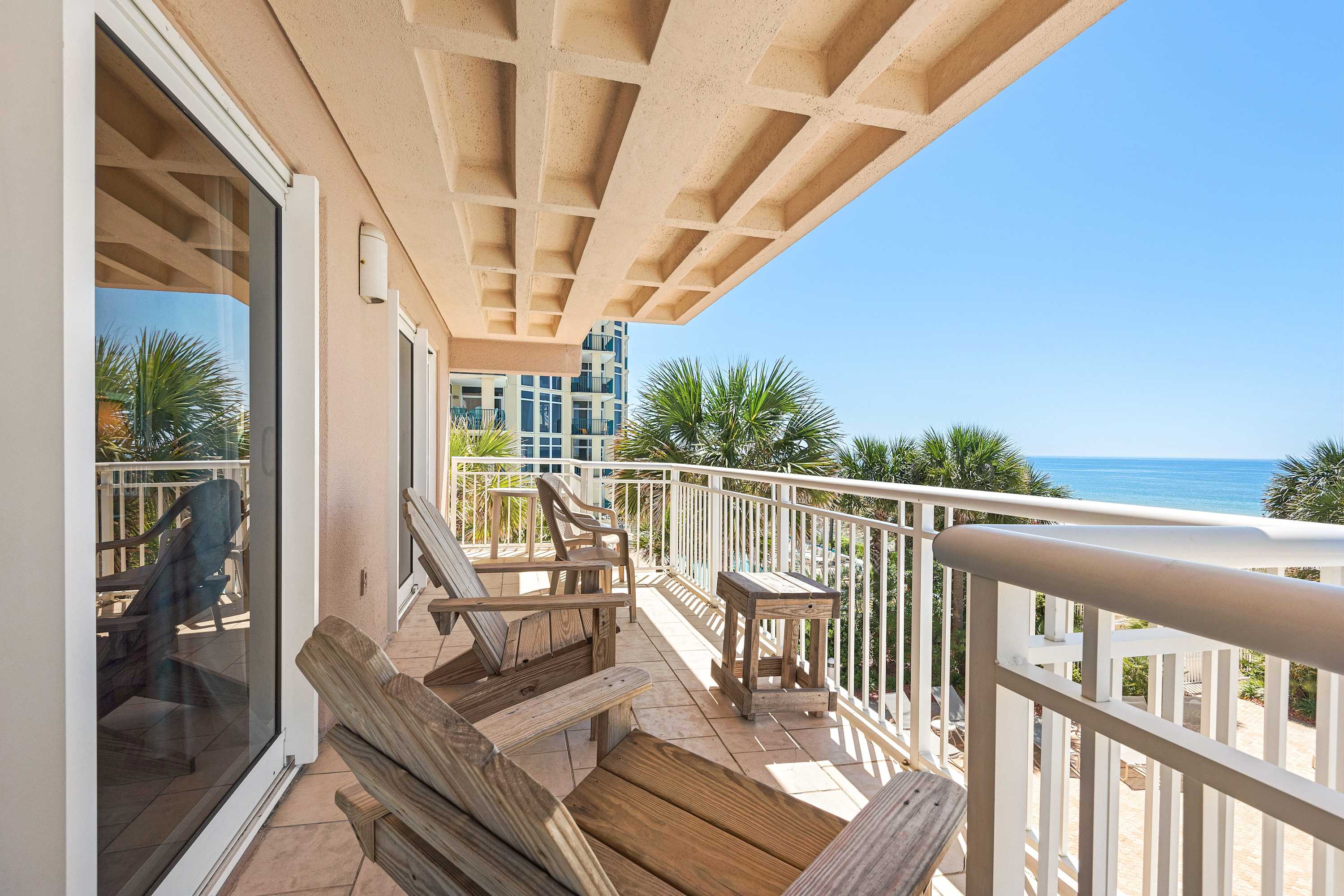 A tiled balcony provides a serene outdoor space with wooden Adirondack chairs and a small side table, offering views of the ocean and lush palm trees from a higher vantage point.
