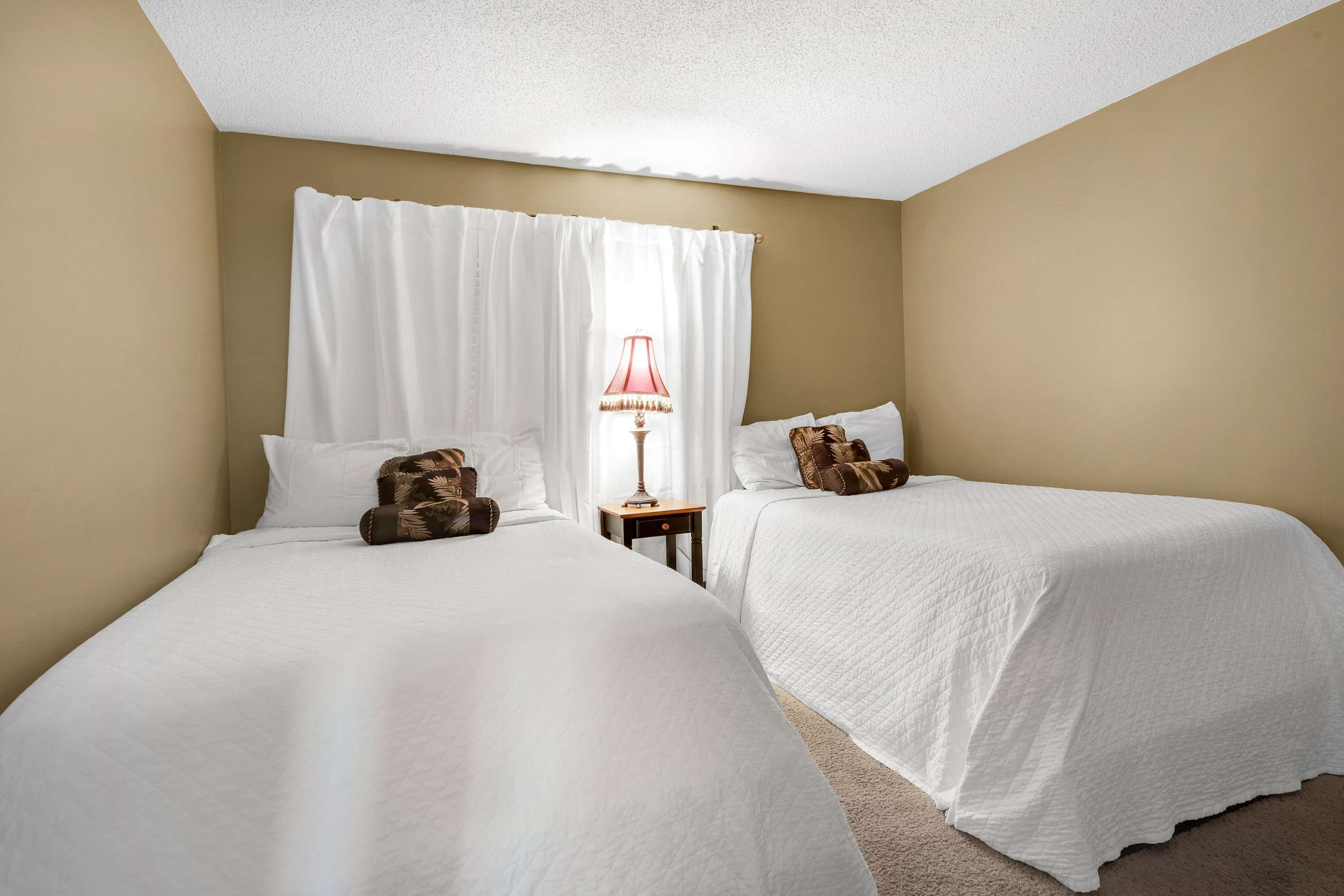 View of a bedroom with two twin beds, each with white bedding and a decorative pillow, separated by a small dark nightstand with a red-shaded lamp. White curtains cover a window.