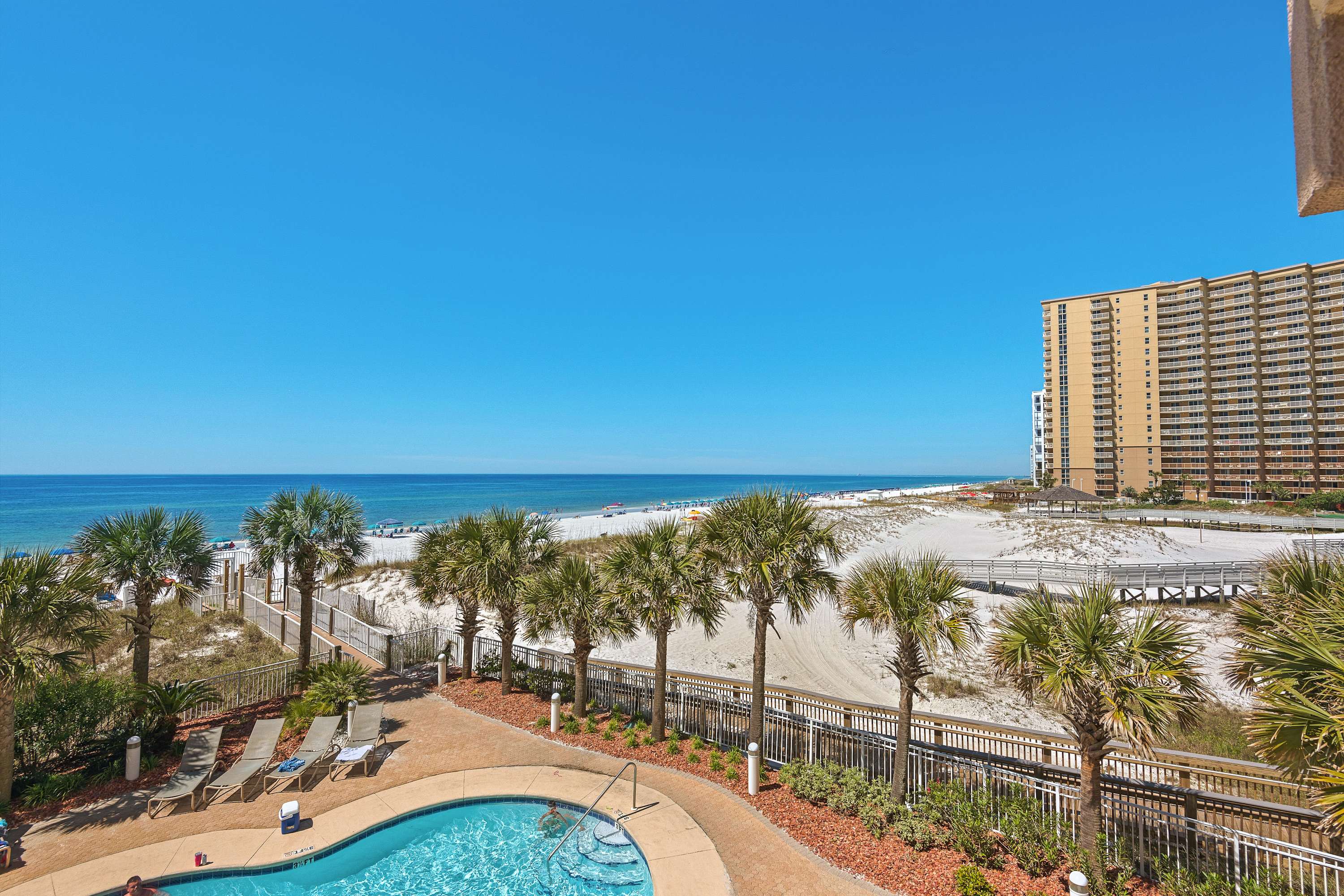 A wide-angle view from the balcony showcases the expansive white sand beach, the clear blue ocean, and a resort pool area lined with palm trees and lounge chairs.