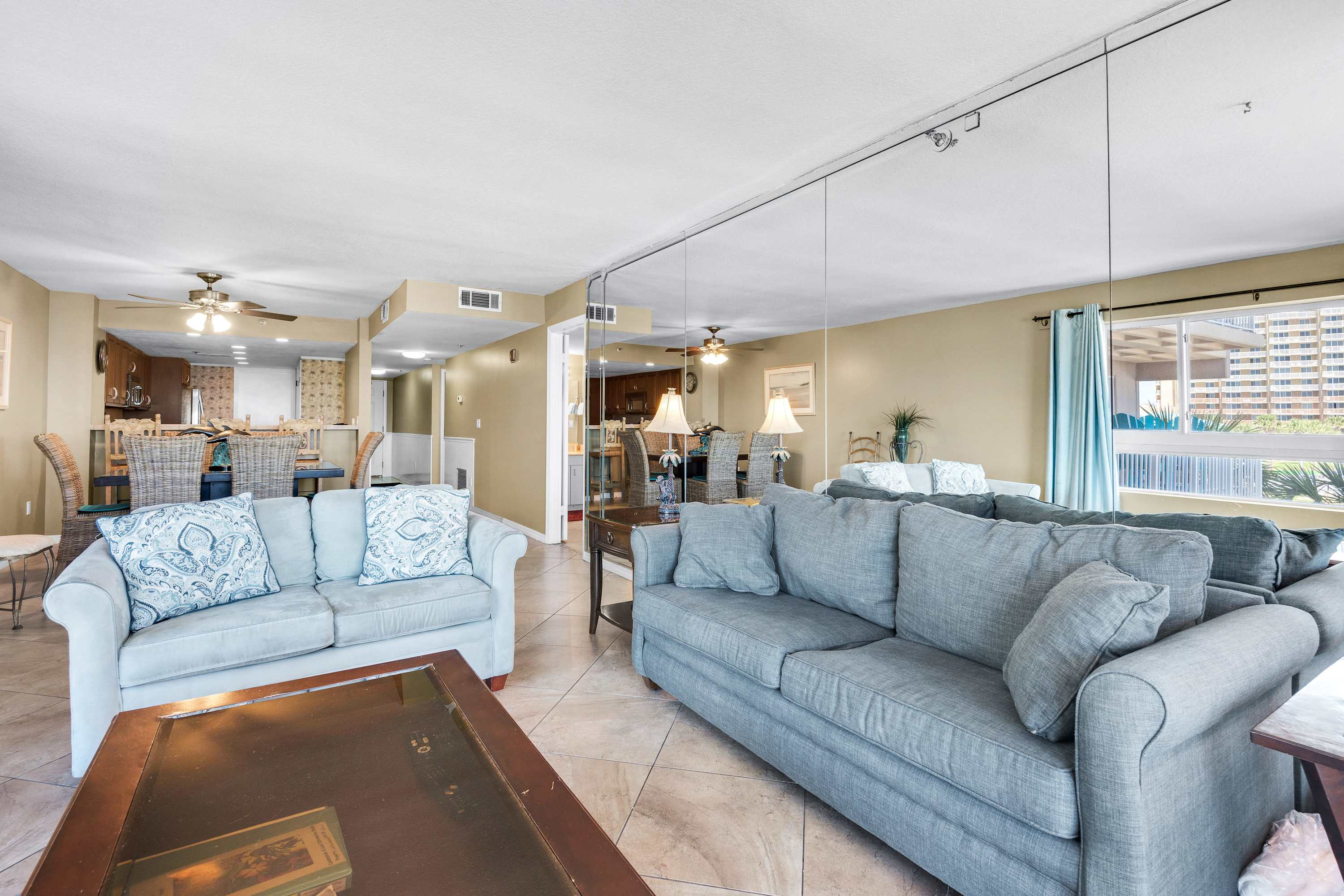 An expansive living area with light-colored walls and tiled flooring, featuring two large sofas and a mirrored wall reflecting the space. The open layout connects to a dining area and kitchen in the background, visible through a pass-through.