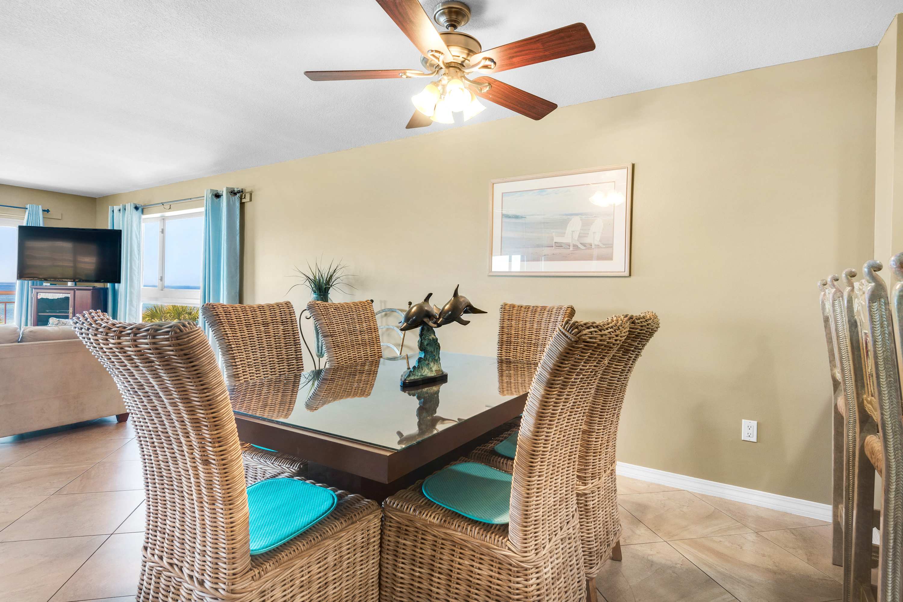 A dining area featuring a square dark wood table with a glass top, surrounded by six wicker chairs with blue cushions, and a decorative dolphin centerpiece. Ocean views are visible in the background.