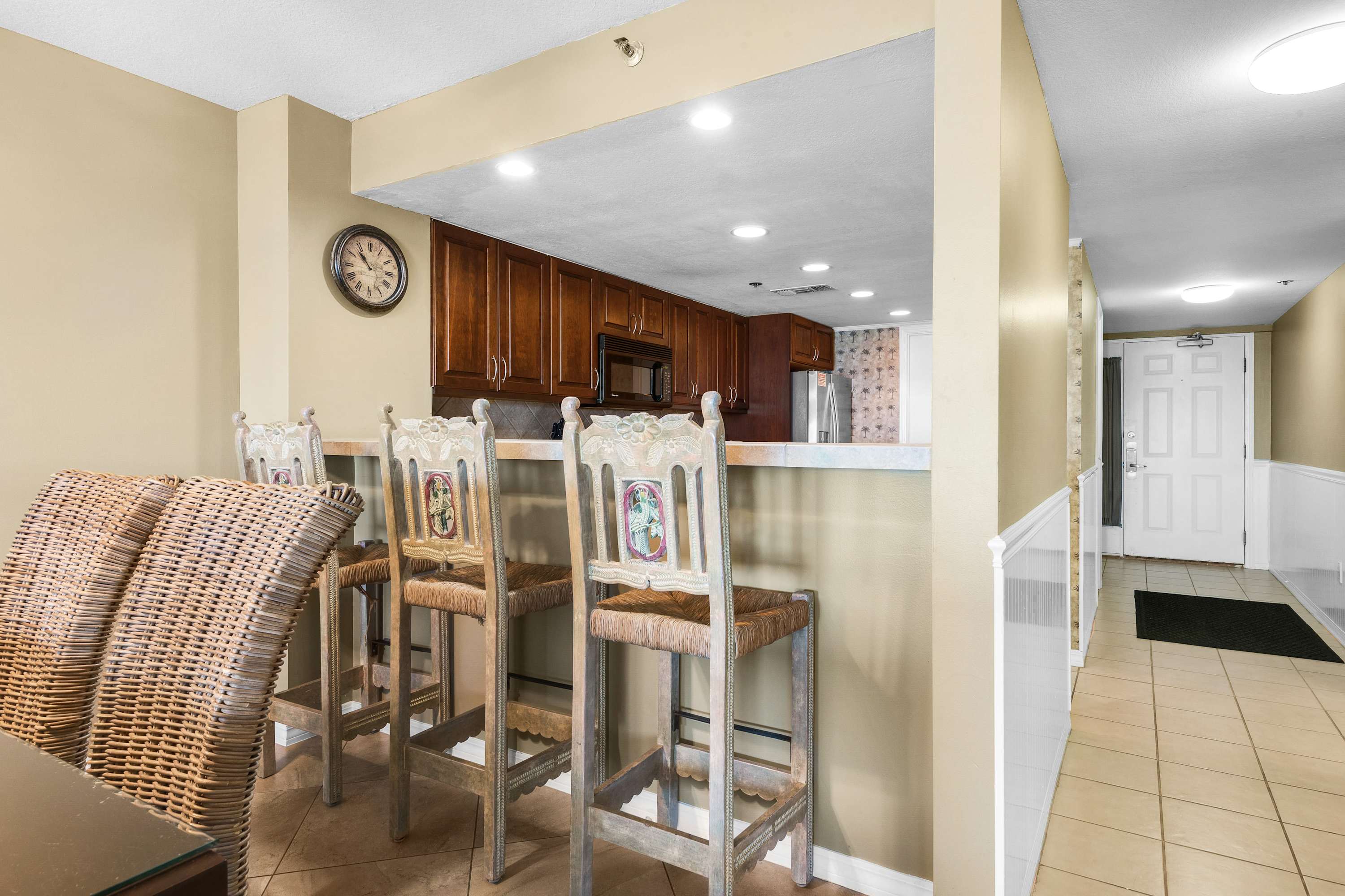 A view from the living area into the kitchen, featuring a breakfast bar with three rustic bar stools with woven seats and decorative backs. The kitchen beyond has dark wood cabinets and light countertops, visible over the bar.
