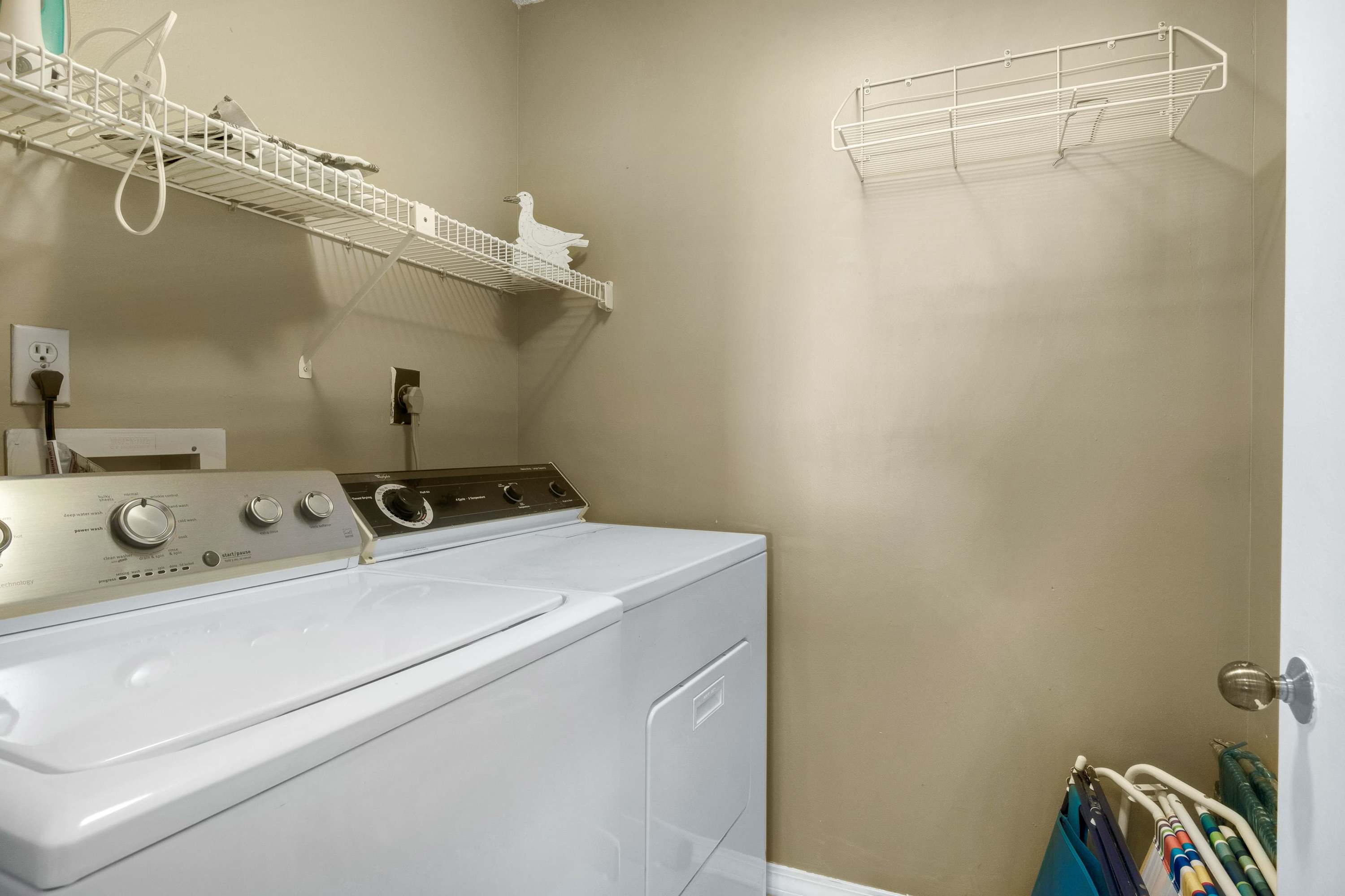 A functional laundry nook features a white top-loading washing machine and a matching dryer, with wire shelving installed above for storage in a neutral-toned room.