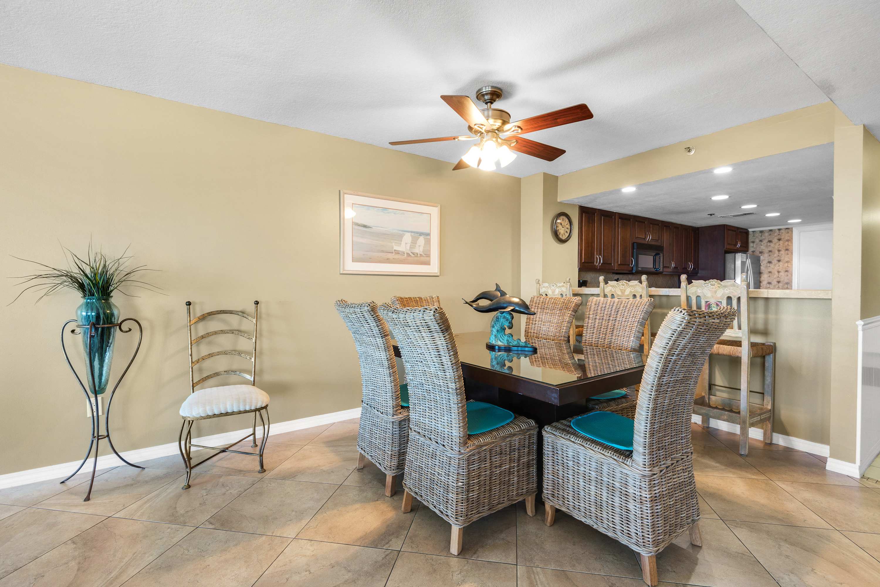 Dining area with a square glass-top table and wicker chairs, adjacent to a kitchen with dark wood cabinets and a breakfast bar, all on light tile flooring.