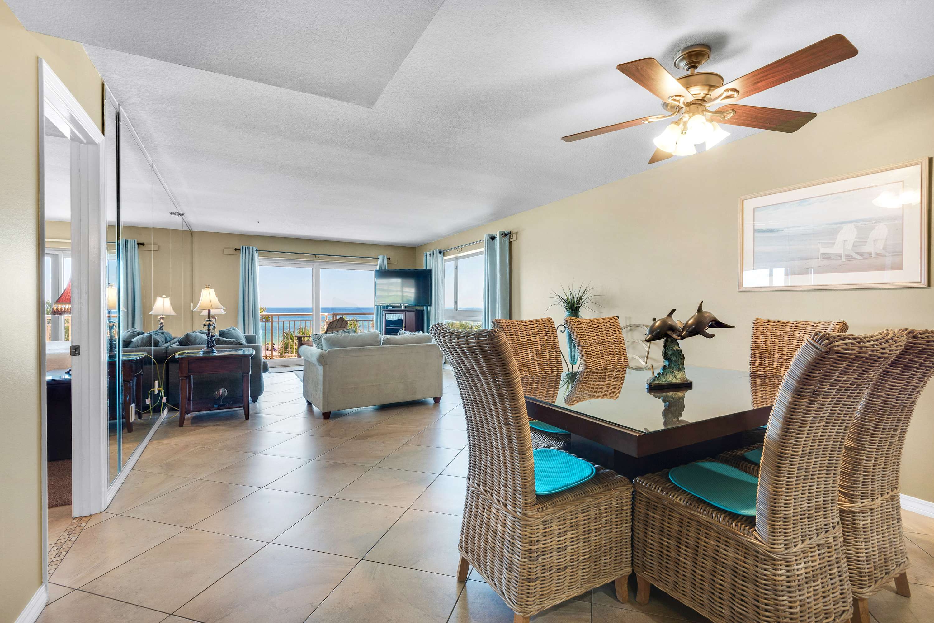 An open-concept living space with a dining area featuring a square table and wicker chairs, leading into a living room with a sofa and panoramic ocean views through large windows.