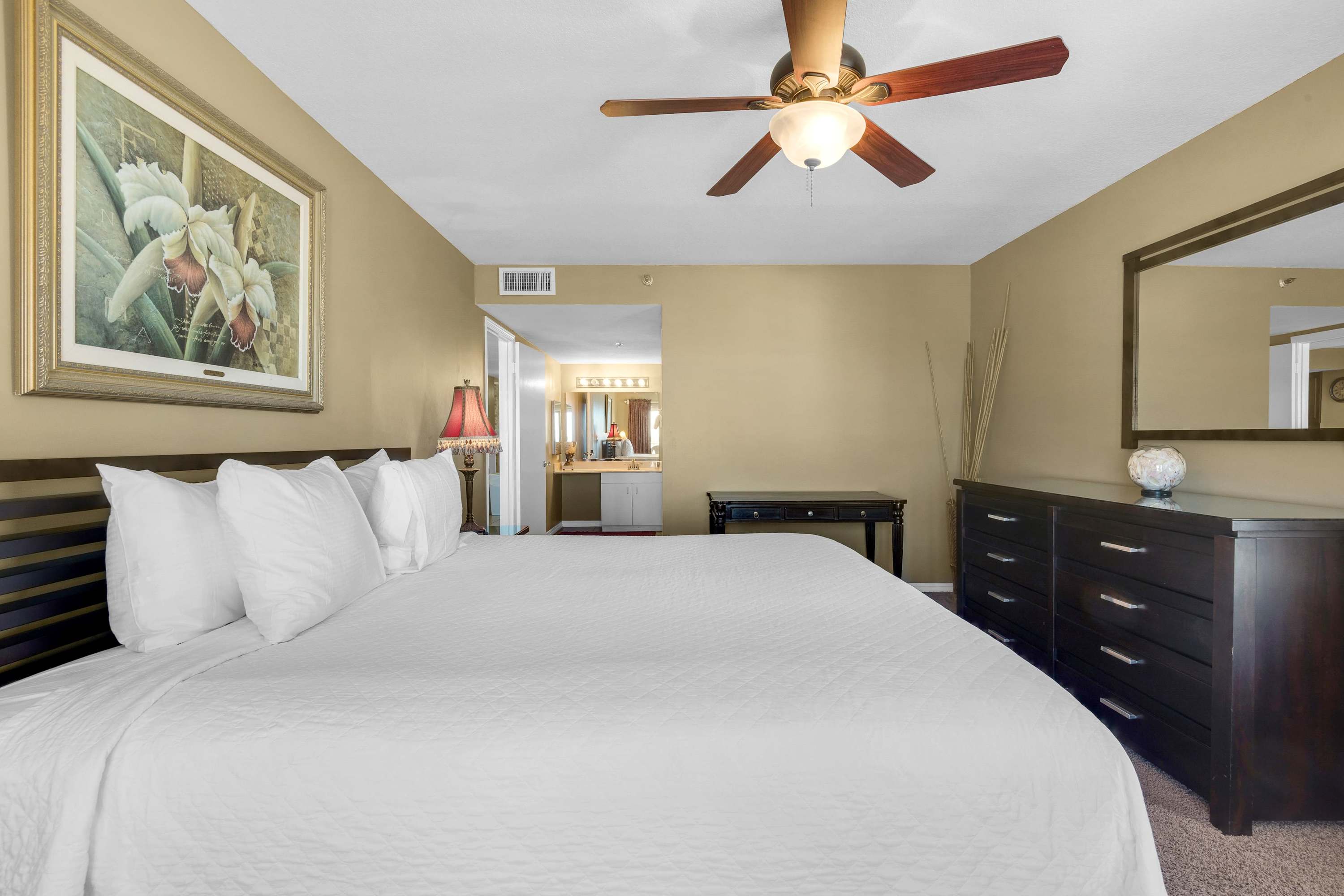 View of the master bedroom with a king bed, dark wood furniture including a dresser and mirror, and a ceiling fan, with a glimpse of the bathroom vanity through an open doorway.