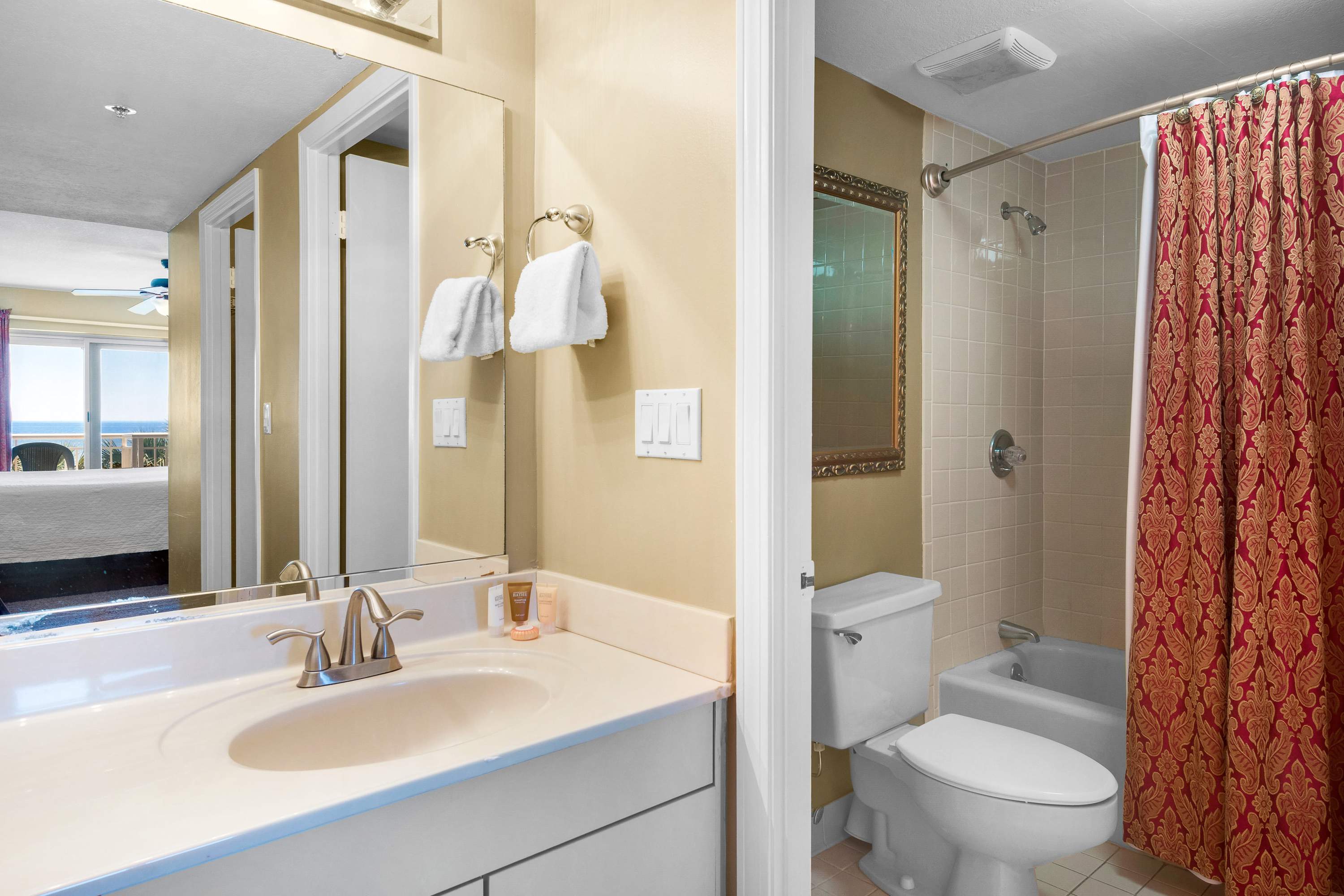 A bathroom featuring a single vanity with a light countertop and chrome faucet, a large mirror, and a toilet and shower/tub combo with a red and gold patterned shower curtain.