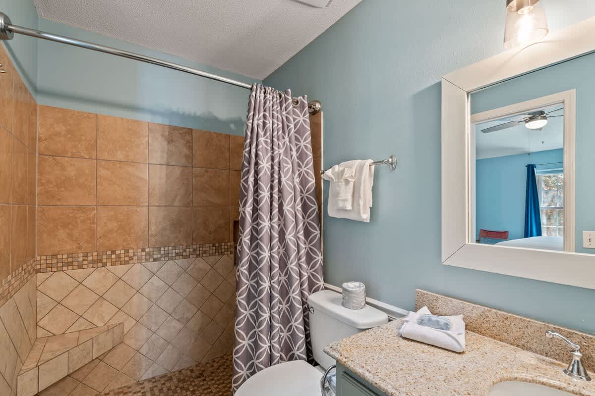 Bathroom featuring light blue walls, a single vanity, and a toilet, with a tiled walk-in shower that includes a built-in bench, enclosed by a patterned shower curtain.