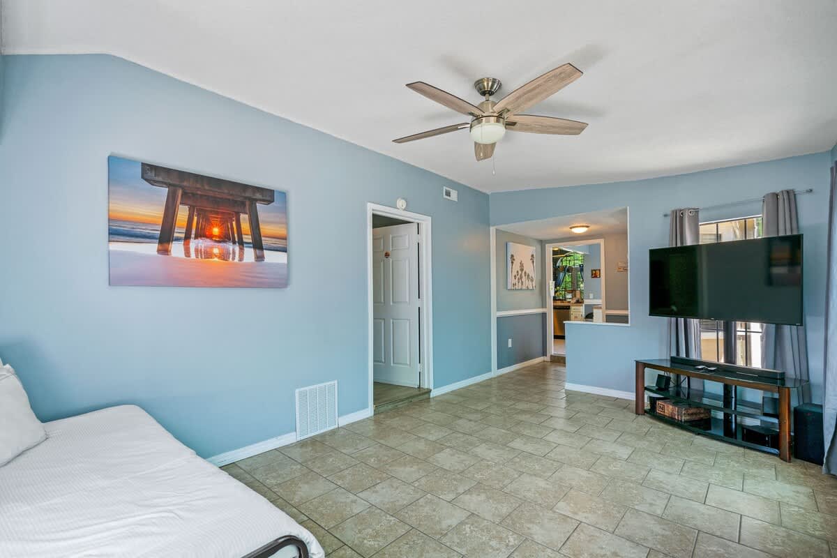 Bright living room with light blue walls and light-colored tile flooring, furnished with a daybed and featuring a ceiling fan, with an open doorway leading to another part of the house.