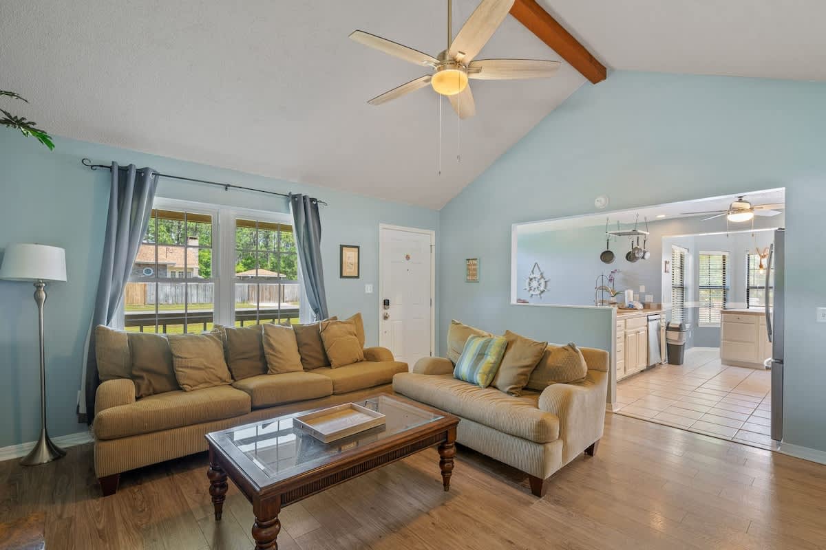 Inviting living room with light blue walls, wood-look flooring, and a vaulted ceiling with a fan, furnished with two large sofas and a glass-top coffee table, offering views of the outdoors through a large window.