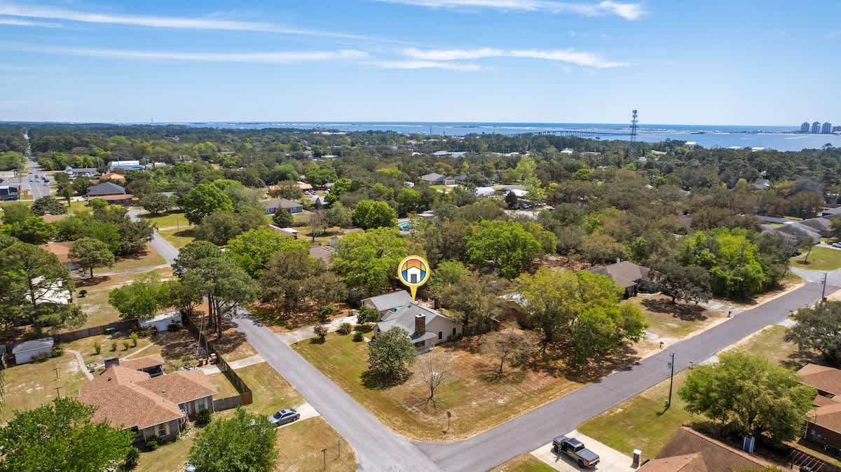 A slightly different aerial perspective of the property, highlighting the corner lot location and the surrounding residential neighborhood with numerous trees. The distant body of water and urban development are still visible on the horizon.