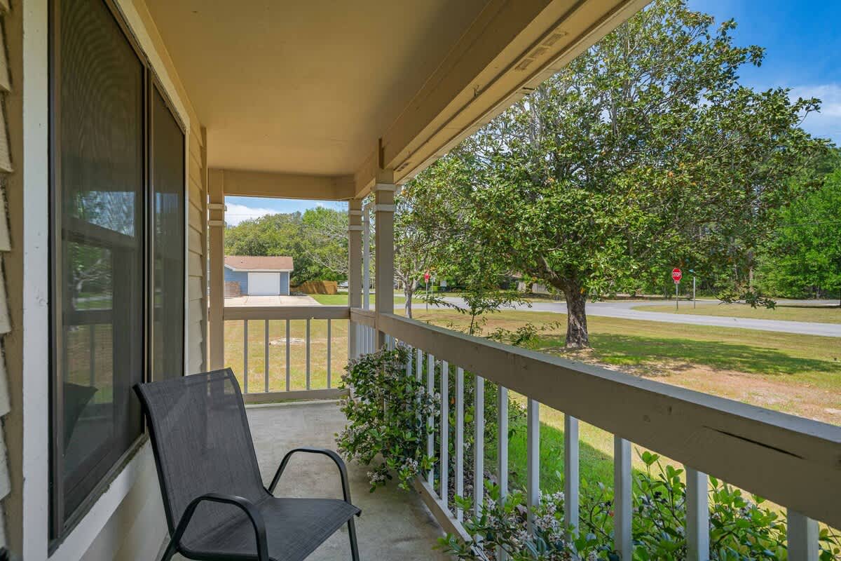 A view from the covered front porch, featuring a dark gray outdoor chair and white railings. Beyond the porch, a large green tree and a residential street with a stop sign are visible under a bright sky.