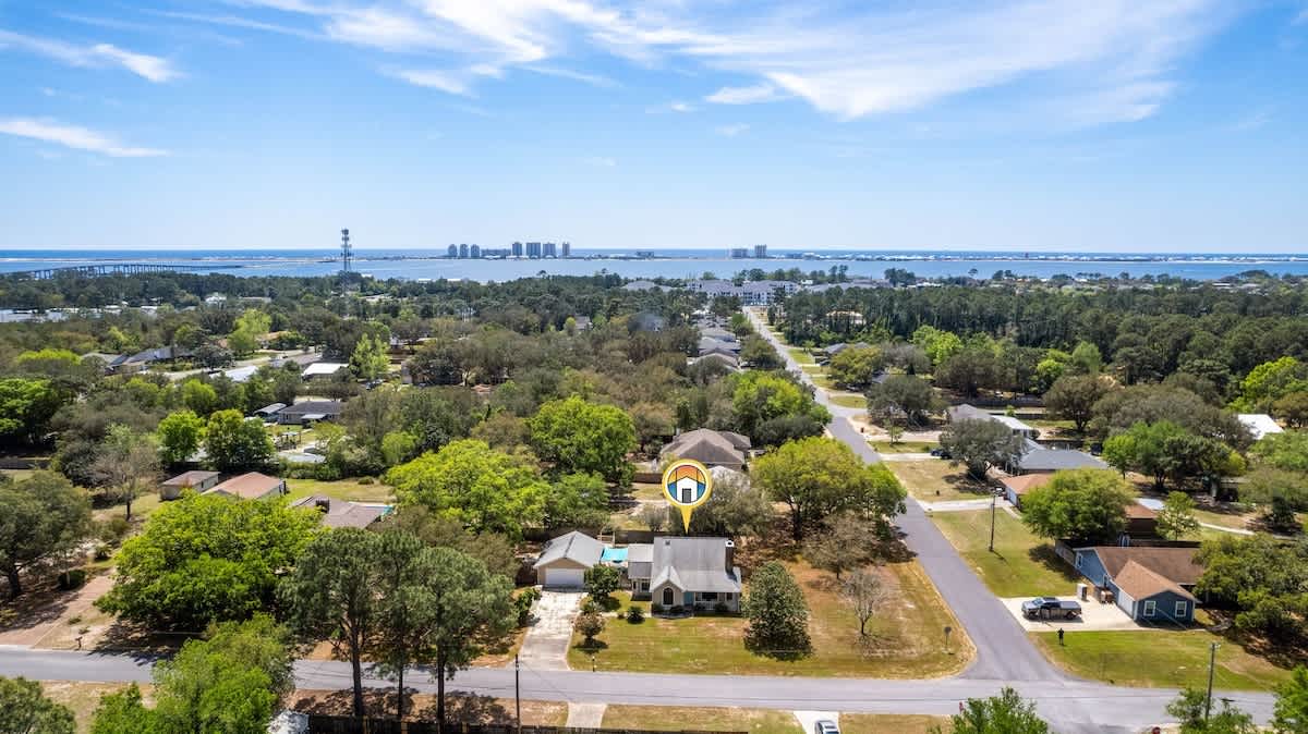 An aerial view of the property, showing the house with its light-colored siding and gray roof, surrounded by a large green yard and mature trees. In the distance, a body of water and a city skyline are visible under a clear blue sky.