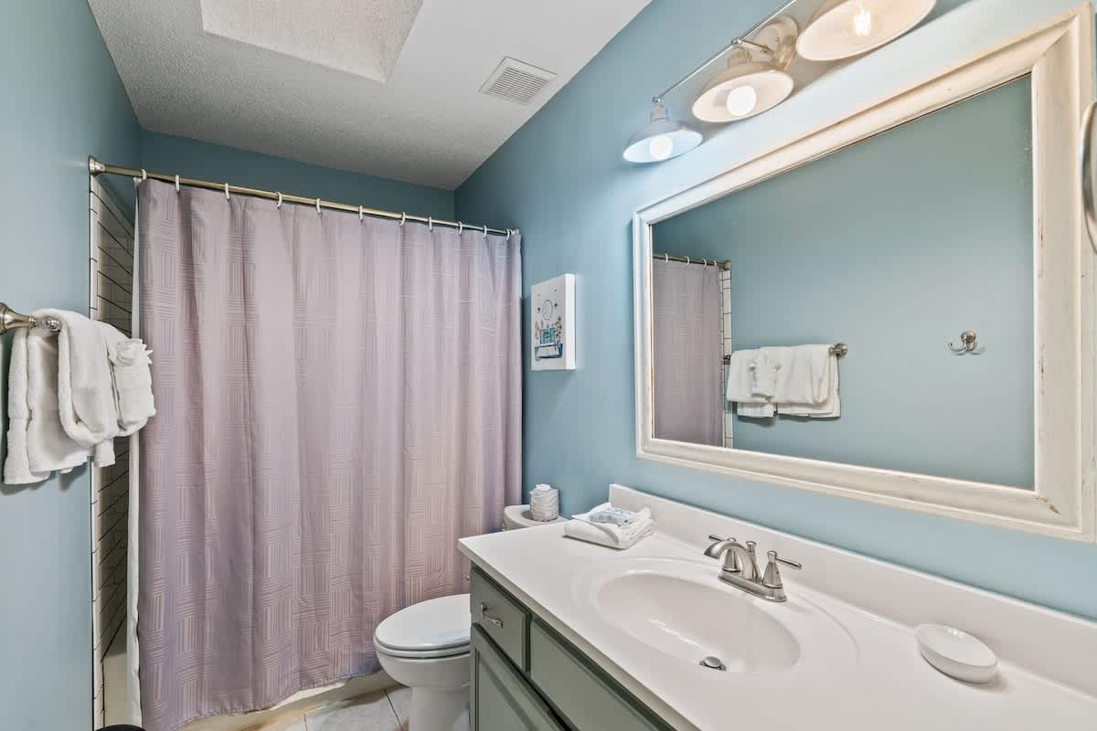 Bathroom with light blue walls, a single vanity with a white countertop, a large framed mirror, and a shower/tub combo with a light purple curtain. White towels are neatly folded on a rack.