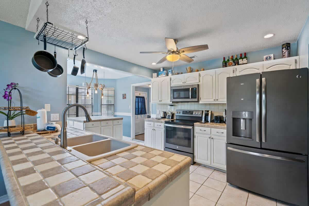 Bright kitchen featuring light blue walls, white cabinetry, and checkered tile countertops, equipped with stainless steel appliances including a refrigerator, microwave, and stove, plus a hanging pot rack.