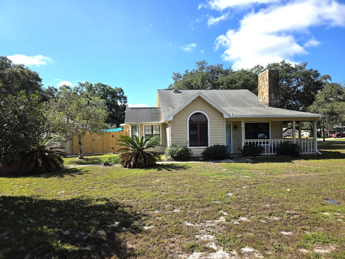 The front of the light yellow house is seen from a slightly different angle, with a large grassy yard in the foreground and mature trees in the background. The sky is blue with white clouds, and a wooden fence is visible to the left of the house.