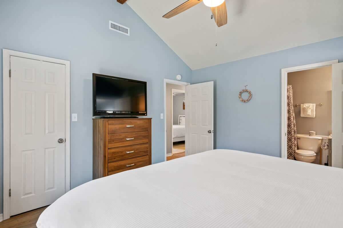 Bedroom featuring a wooden bed frame with white linens, light blue walls, and wood-look flooring, equipped with a dresser and television, with an open door revealing the adjacent bathroom.