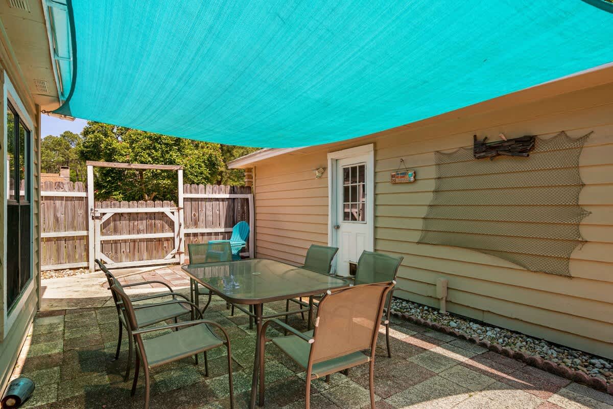 A paved patio area features a glass-top dining table with six chairs, shaded by a large turquoise sunshade. The area is adjacent to a light-colored house wall with a white door and a wooden fence in the background.