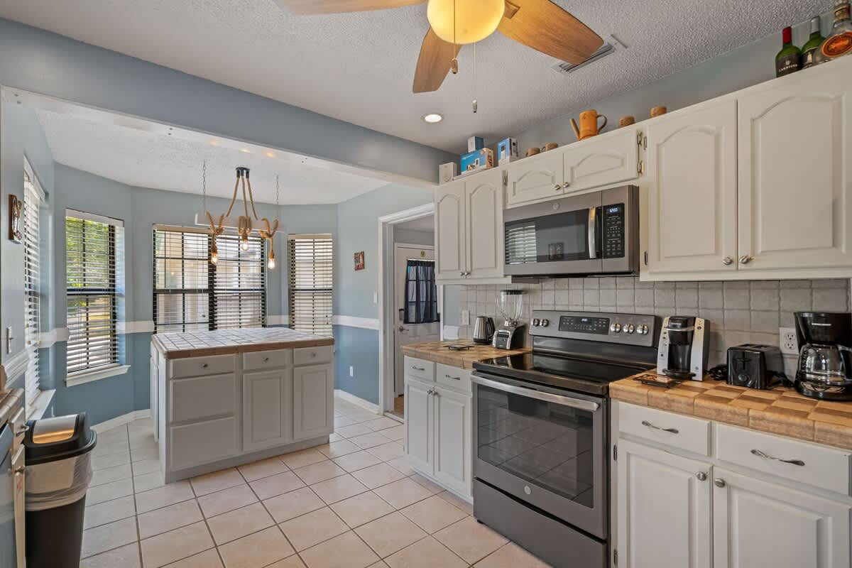 Kitchen area showcasing white upper and lower cabinets, checkered tile countertops, and stainless steel appliances including an over-the-range microwave and electric stove, with a kitchen island visible in the background.