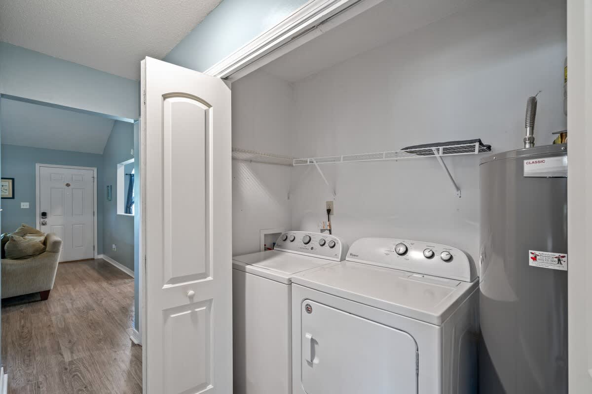 Compact laundry closet featuring a white top-loading washing machine and a front-loading dryer, with a water heater and wire shelving above. A doorway to a living area is visible.
