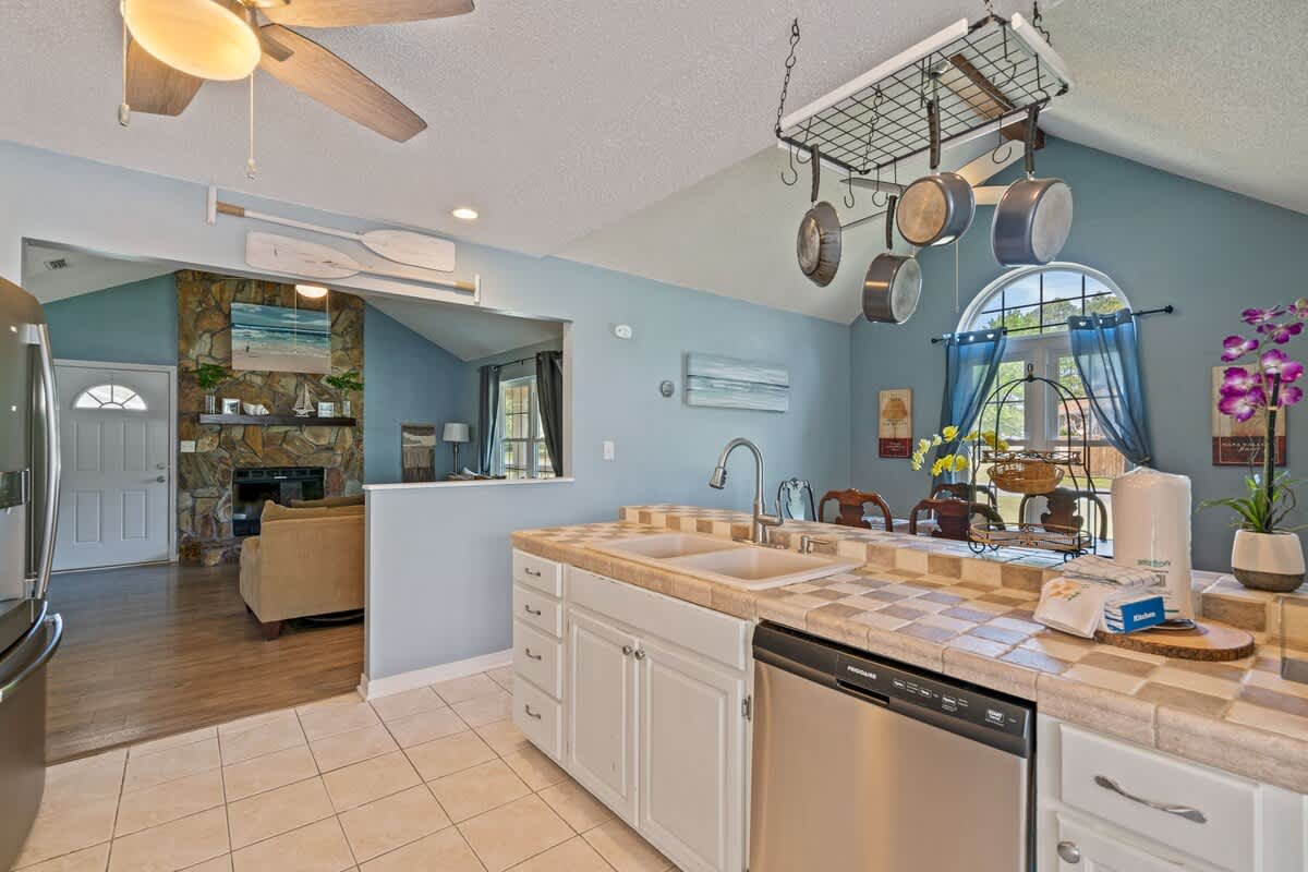 Functional kitchen area with white cabinets, tiled countertops, and a double sink, featuring a pot rack hanging above, with a pass-through window offering a view into the living room.